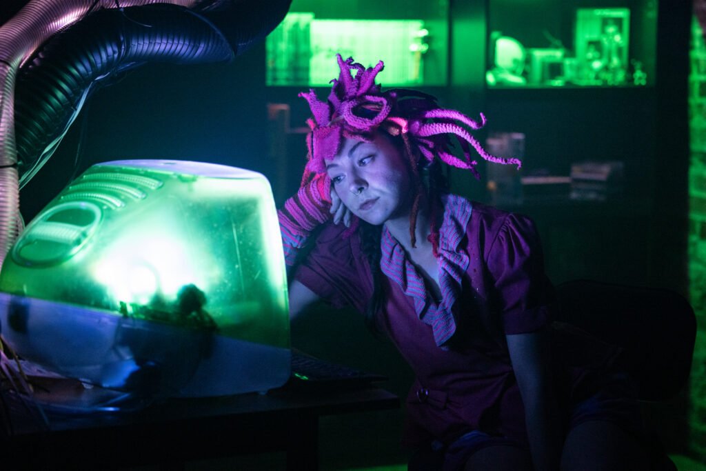 A woman with pink dreadlocks, wearing a maroon shirt and purple scarf, sits in a dimly lit room illuminated by green and purple lights. She looks tired or bored as she leans her head on her hand, facing an old green computer monitor.