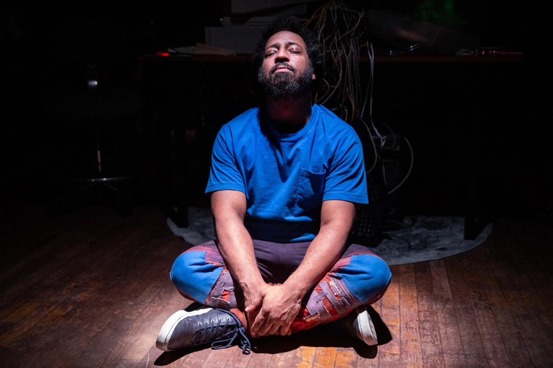 A man in a blue shirt and ripped pants sitting cross-legged on a wooden floor with his eyes closed, illuminated by a spotlight in a dark room.