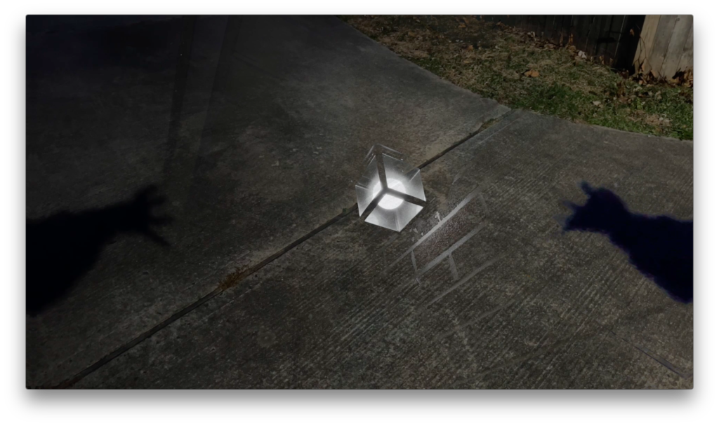 An outdoor scene featuring a concrete sidewalk at night, with a small illuminated cube-shaped lamp placed on the ground casting reflections and shadows, including a silhouette of a horse figure on the right side.