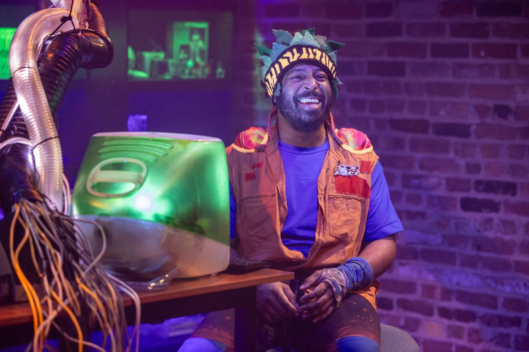 A man wearing a colorful beanie and a bright orange vest sitting at a table with a vintage computer, smiling in a room with purple and green lighting and a brick wall background.