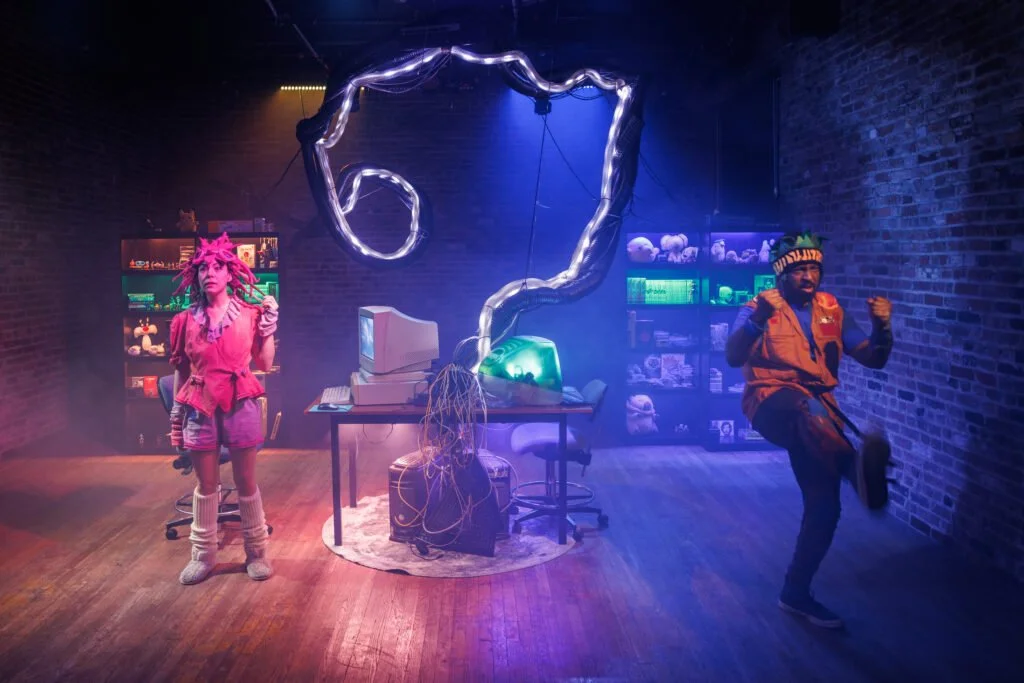 Colorful scene with two people in costumes, one wearing a pink outfit with a pink wig and another in a brown jacket with a green hat, in a room with neon lights, a computer, and shelves filled with stuffed animals. The room has a dark, industrial aes