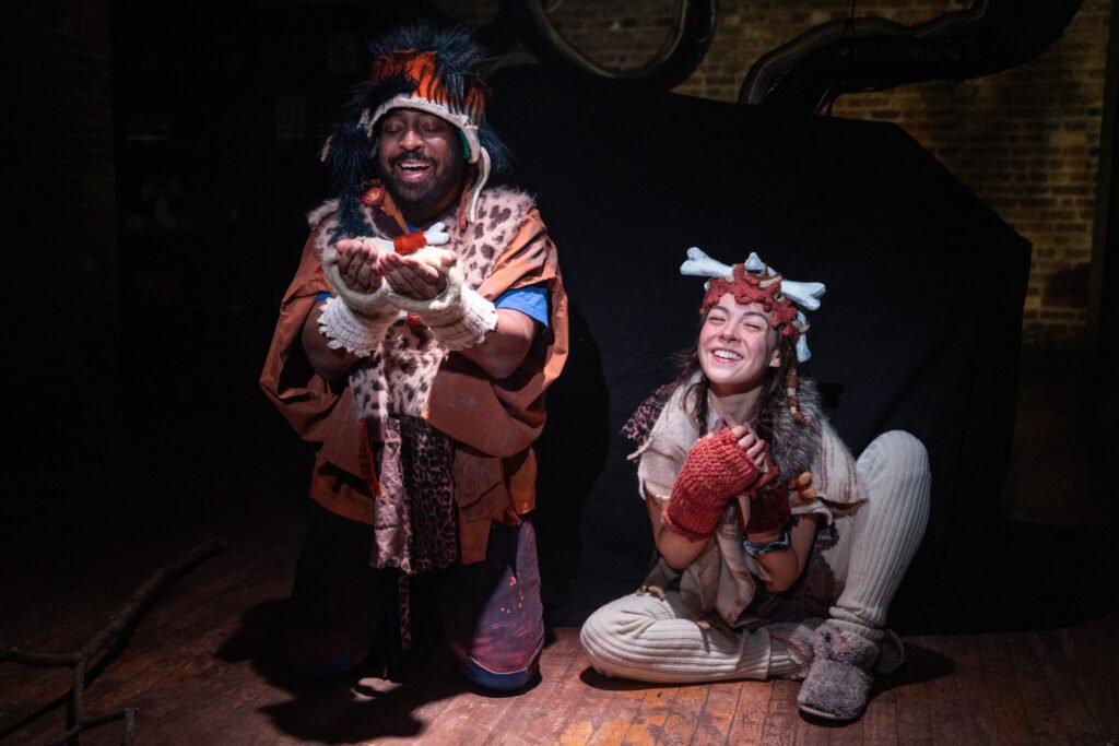 Two people in costume with theatrical makeup and accessories, one standing and holding a small object, the other sitting on the ground smiling, in an indoor setting with dark background.
