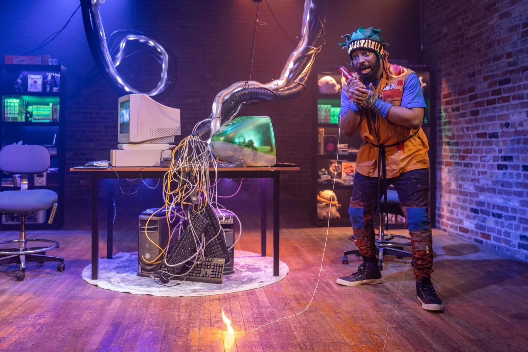 A man in a colorful, eccentric outfit with a multicolored hat and colorful pants stands in a room with exposed brick walls, surrounded by old computer equipment, wires, and electronic components. He appears excited or surprised, holding a small objec