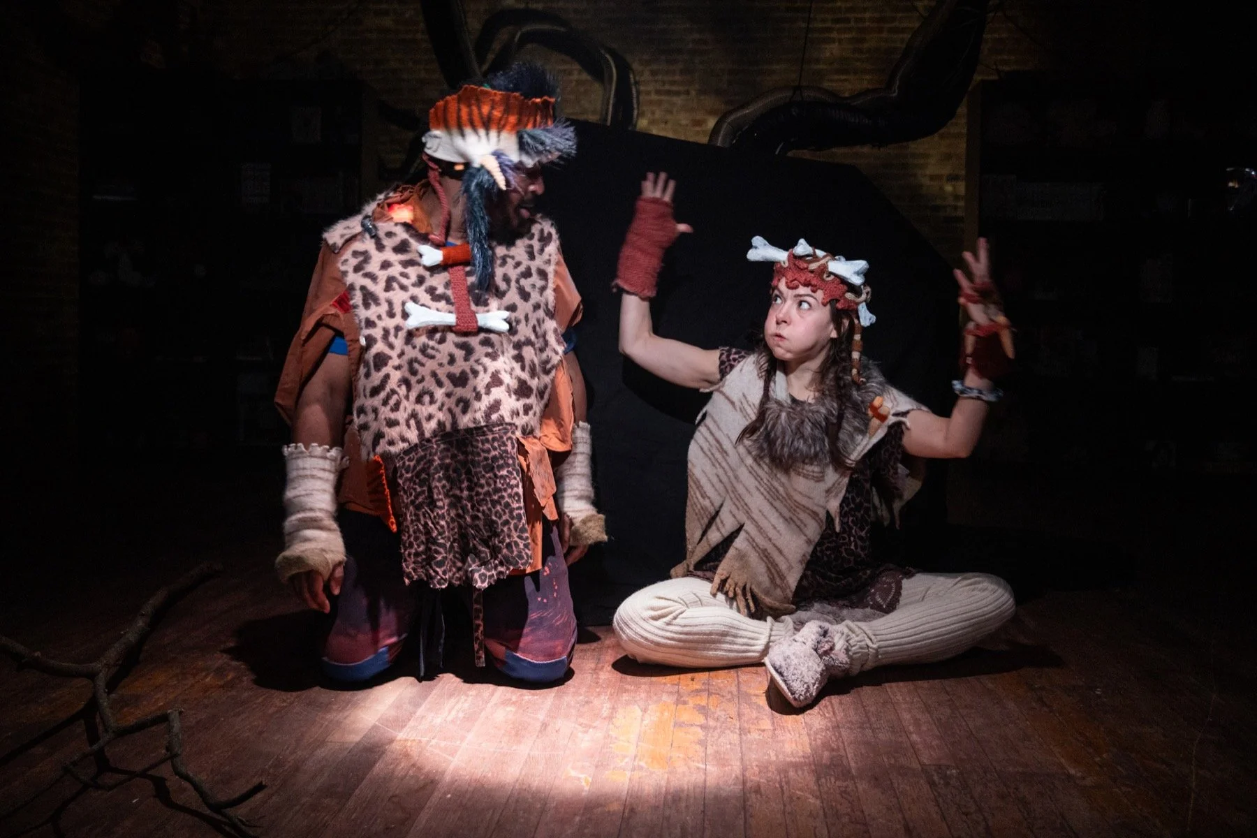 Two actors dressed as cavemen performing on stage, with one standing and the other sitting cross-legged. Both are wearing animal print costumes and accessories, with expressive poses against a dark background.