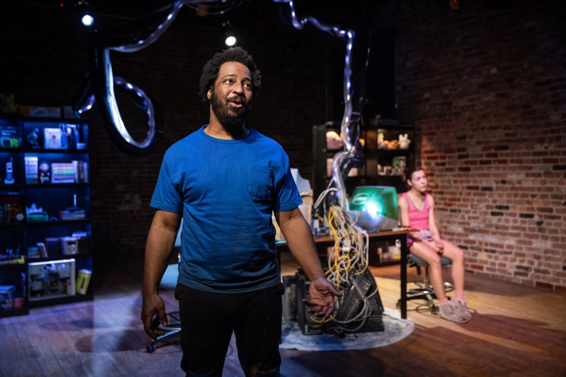 Two people in a room with brick walls, one man in a blue shirt standing in the foreground, a woman in a pink top sitting on a chair in the background, with electronic equipment and books around.