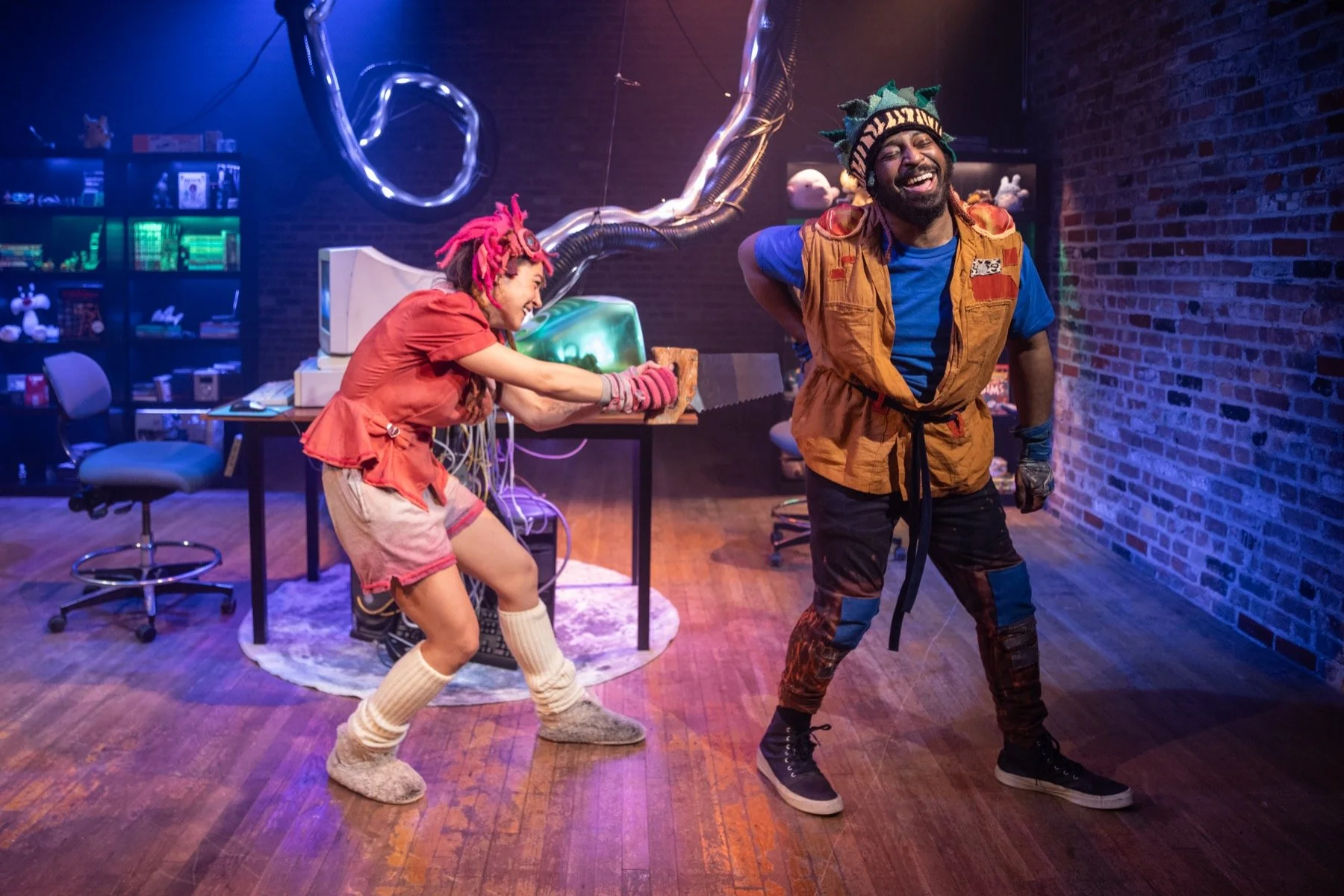 Two people, one with pink dreadlocks and another with a colorful hat, are laughing and playing in a room with a brick wall and various electronic equipment.