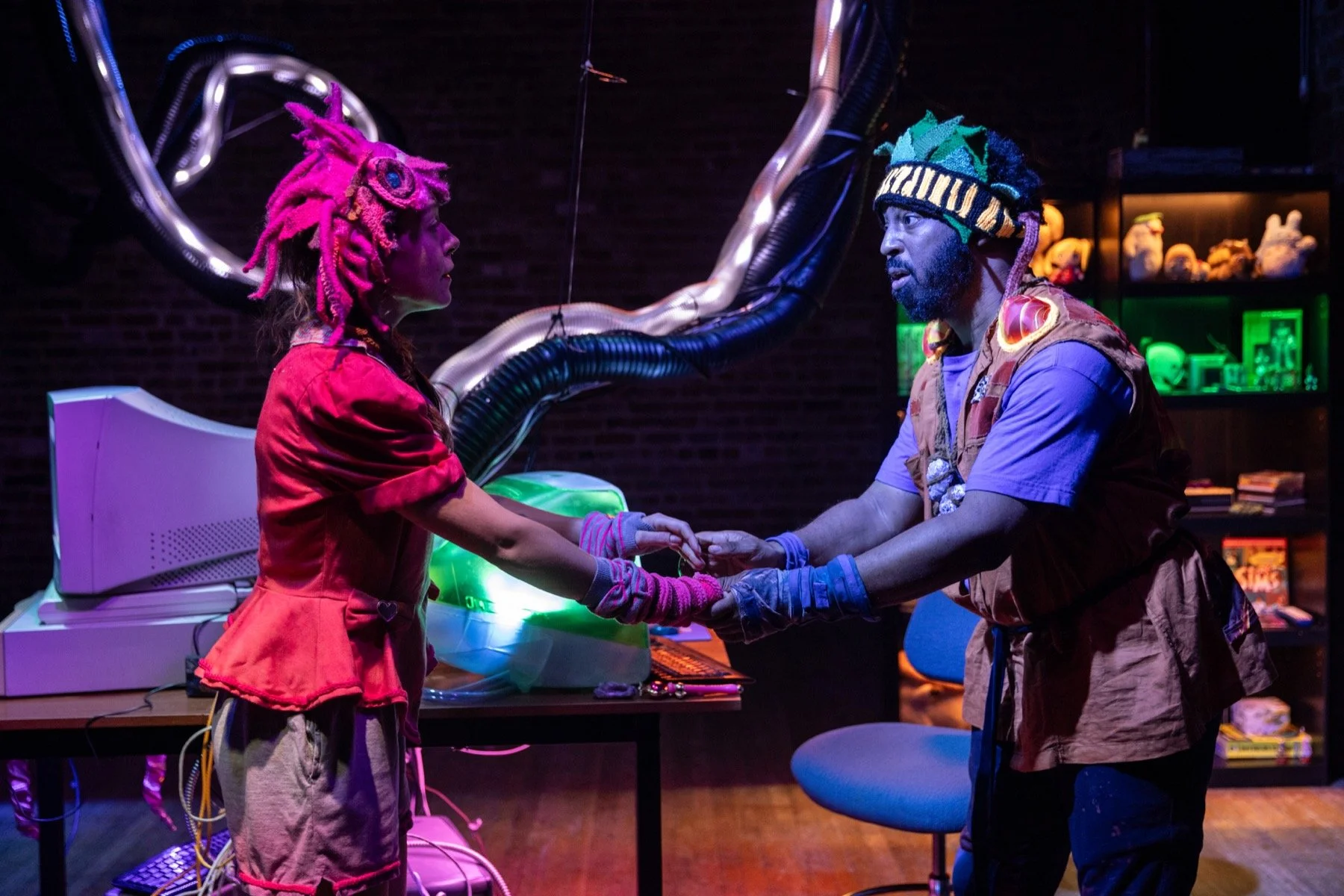 Two people in colorful costumes holding hands in a dimly lit room with sci-fi decor and computer equipment.