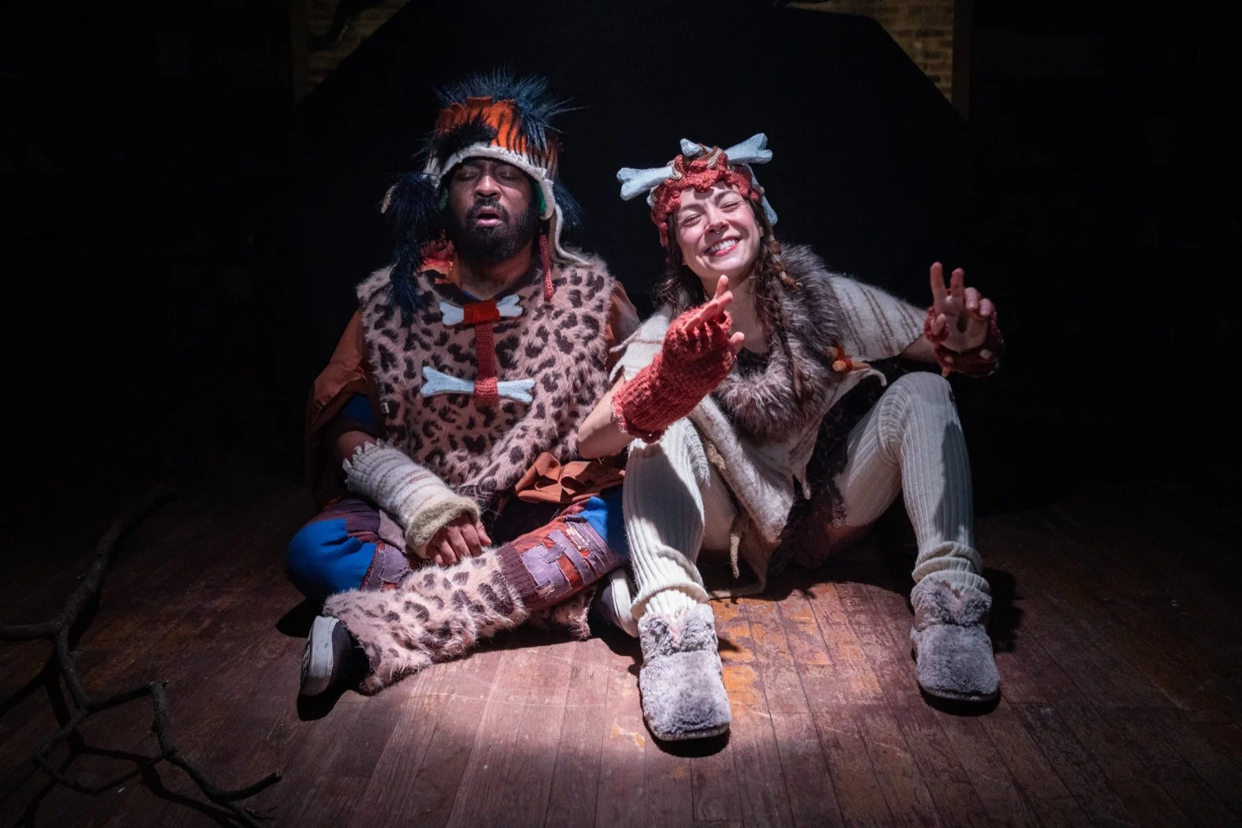 Two actors on stage in costume, one dressed as a caveman with a leopard print outfit, and the other as a whimsical character with a headpiece and knitwear, sitting on a wooden floor under spotlight.