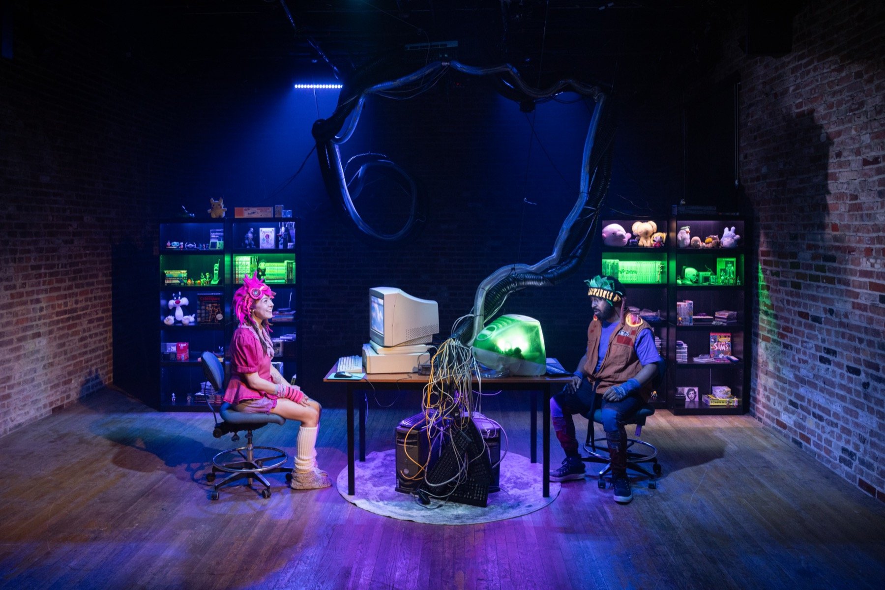 A theatrical scene with two people in a room with brick walls and wooden floors. The person on the left has pink hair and clothing, while the person on the right wears a virtual reality headset and casual attire. The center has vintage computer equip