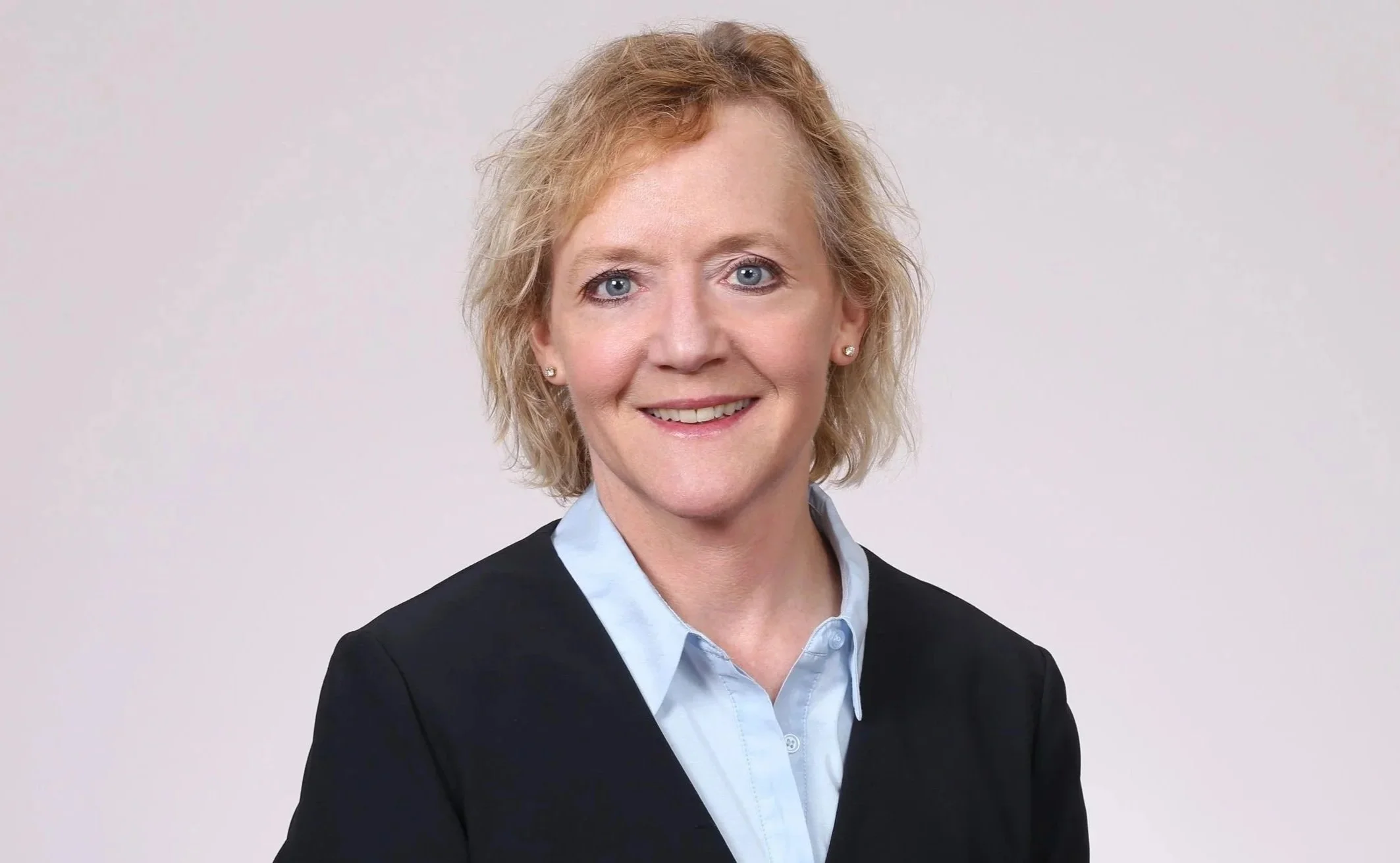 A professional woman with short, curly blonde hair wearing a black blazer and light blue collared shirt, smiling against a plain white background.