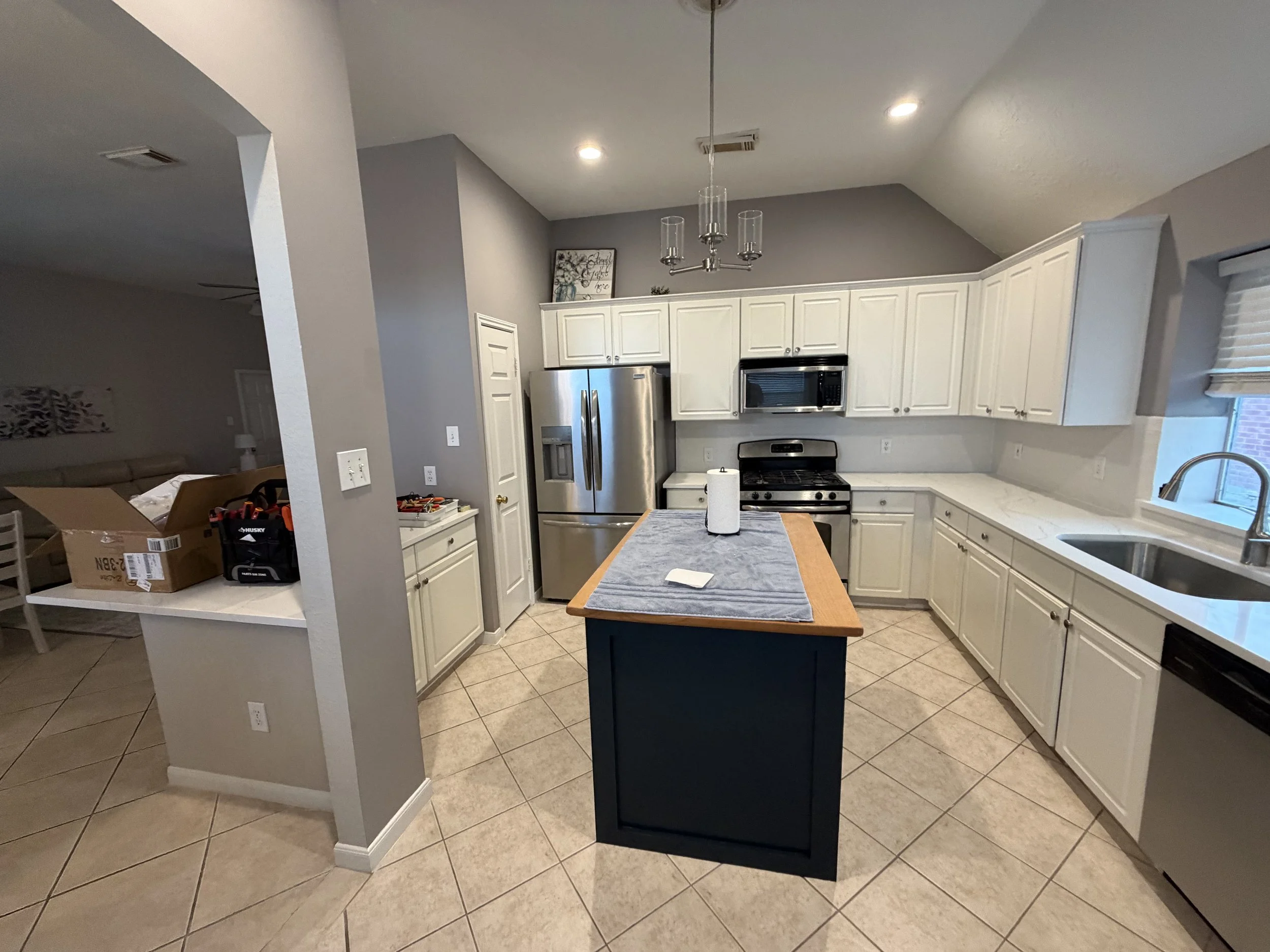 Kitchen with white cabinets, stainless steel refrigerator, stove, microwave, granite countertops, and a small island with a paper towel roll. There is a window with blinds and a ceiling light fixture. Items in the foreground include a cardboard box a