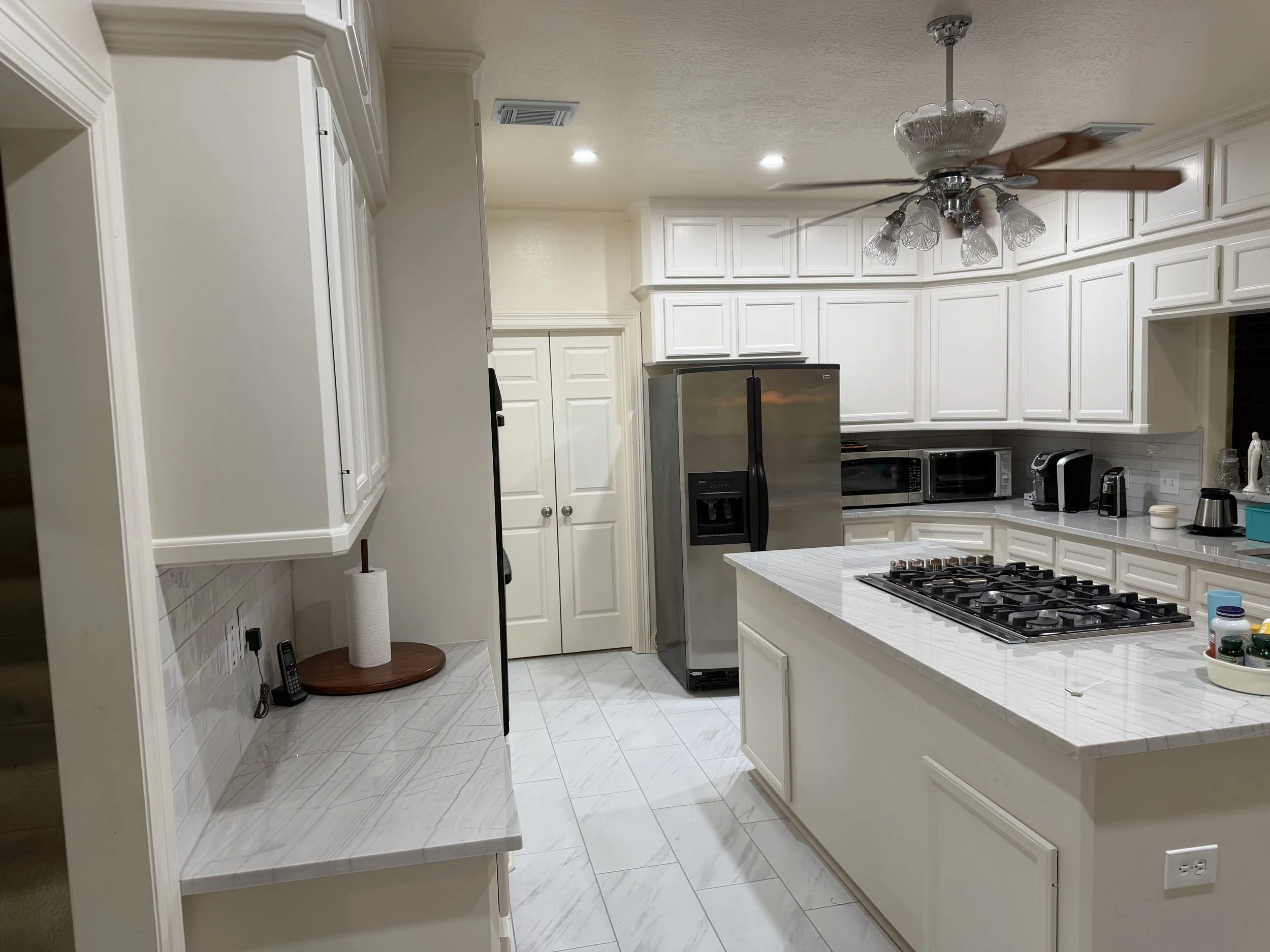 Kitchen with white cabinets, a stainless steel refrigerator, a gas stove, microwave, coffee maker, and several containers on the counter. Ceiling fan with glass light fixtures and recessed lighting. White tiled floor and a large island with a white m