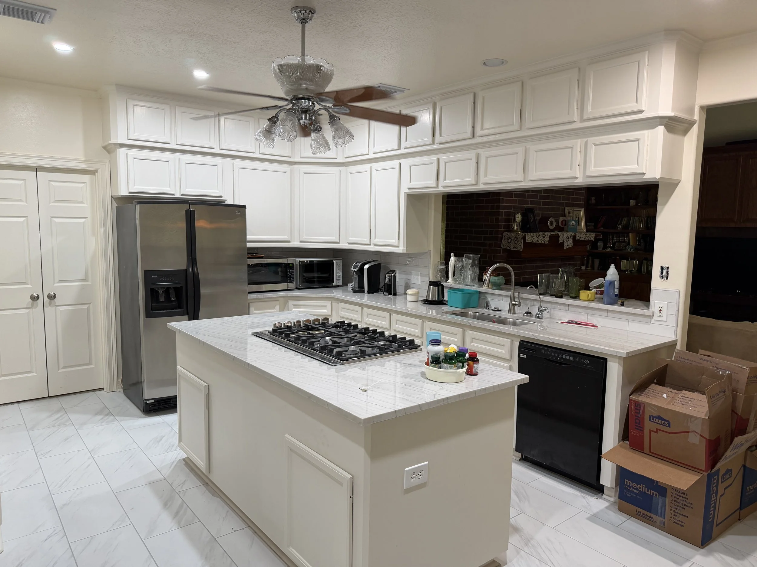 A kitchen with white cabinets, a kitchen island, stainless steel refrigerator, microwave, and various small appliances. There are also boxes and storage items nearby.