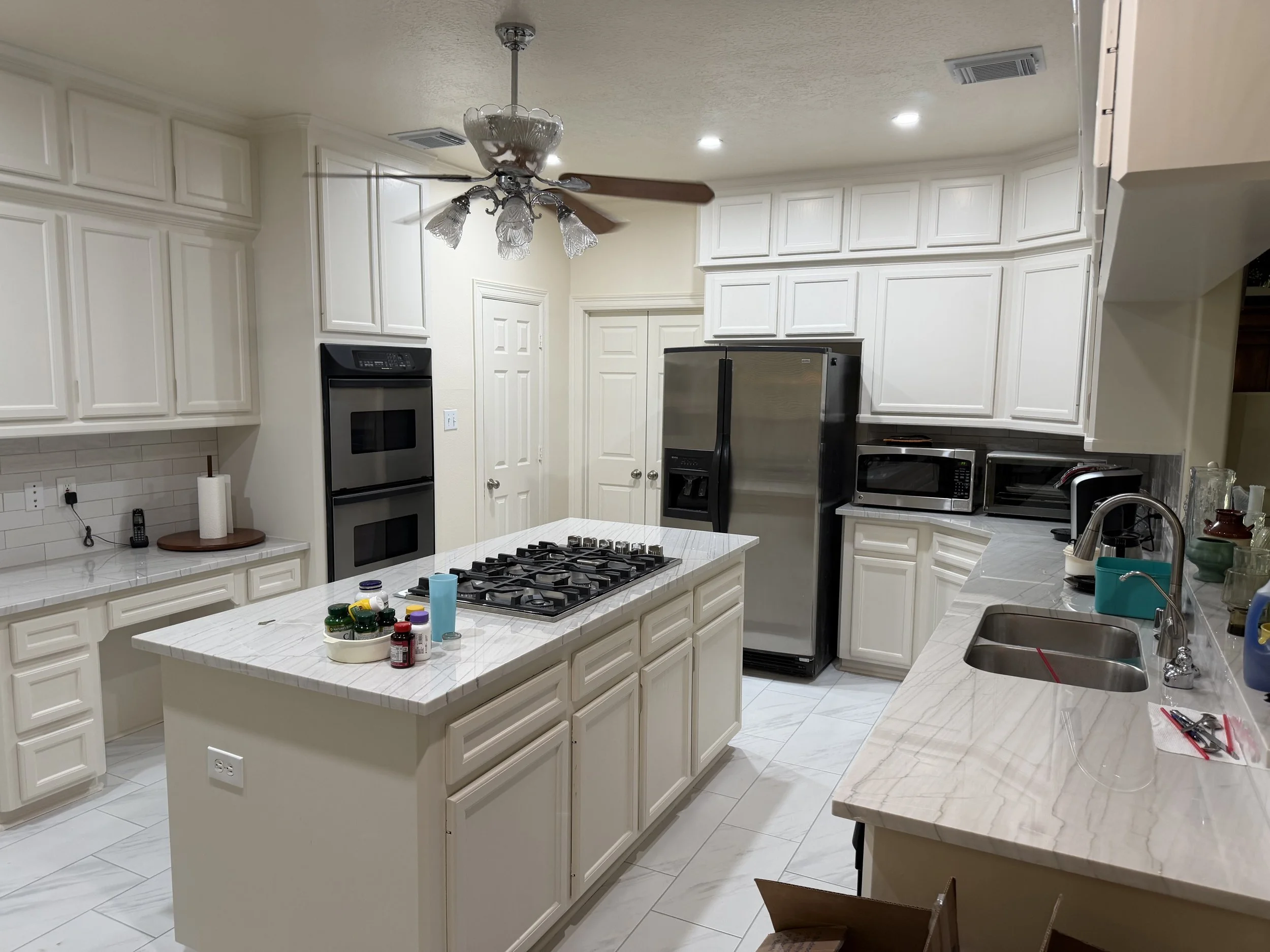 A spacious white kitchen with white cabinets, marble countertops, and stainless steel appliances including a double oven, refrigerator, microwave, and white cabinets. An island with a stove is in the center, and various food and kitchen items are on 