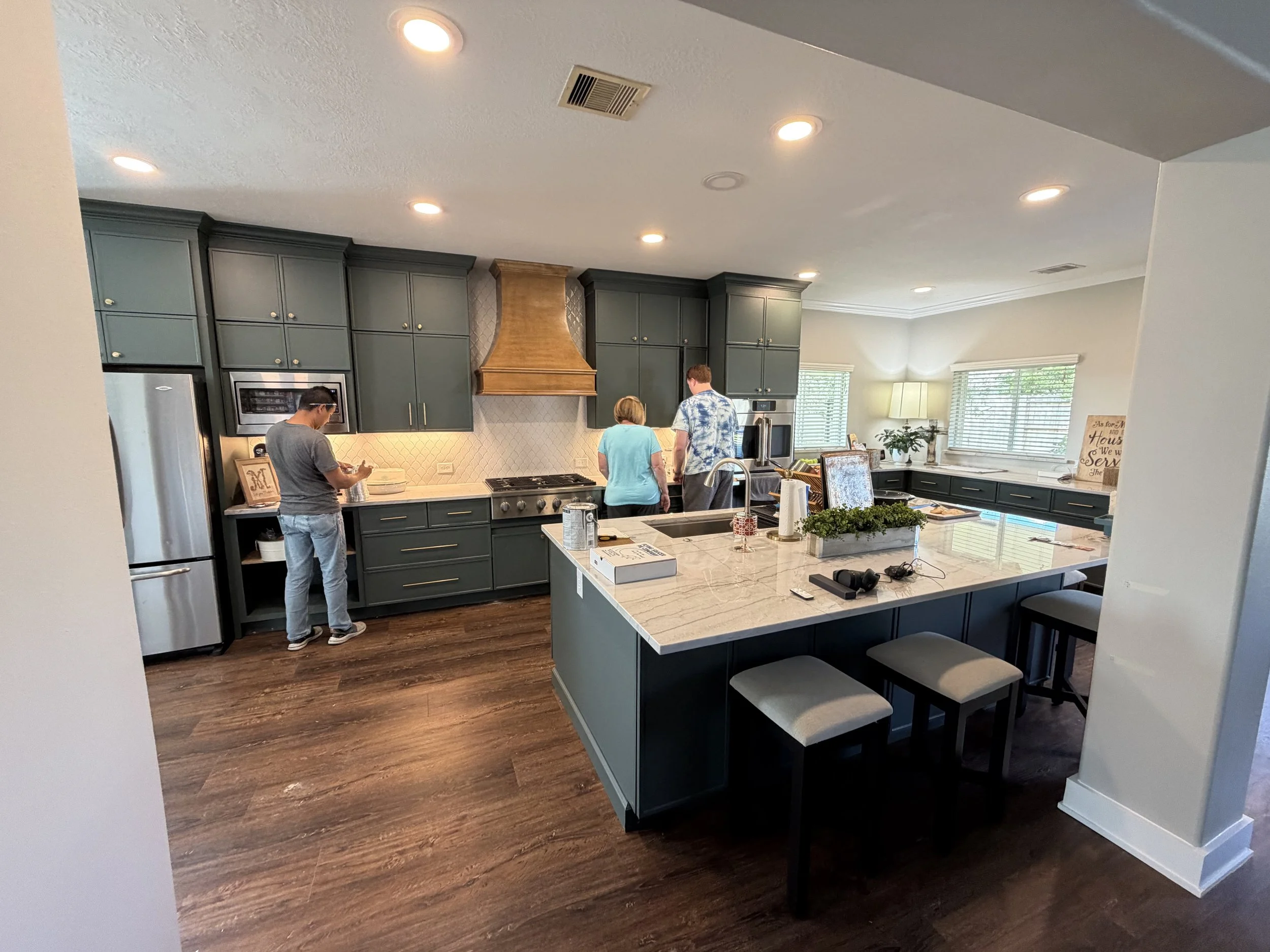 A modern kitchen with dark green cabinets, a large white marble island, stainless steel appliances, a brown range hood, and three people gathered around the counter. There are windows with white blinds and a small indoor plant.