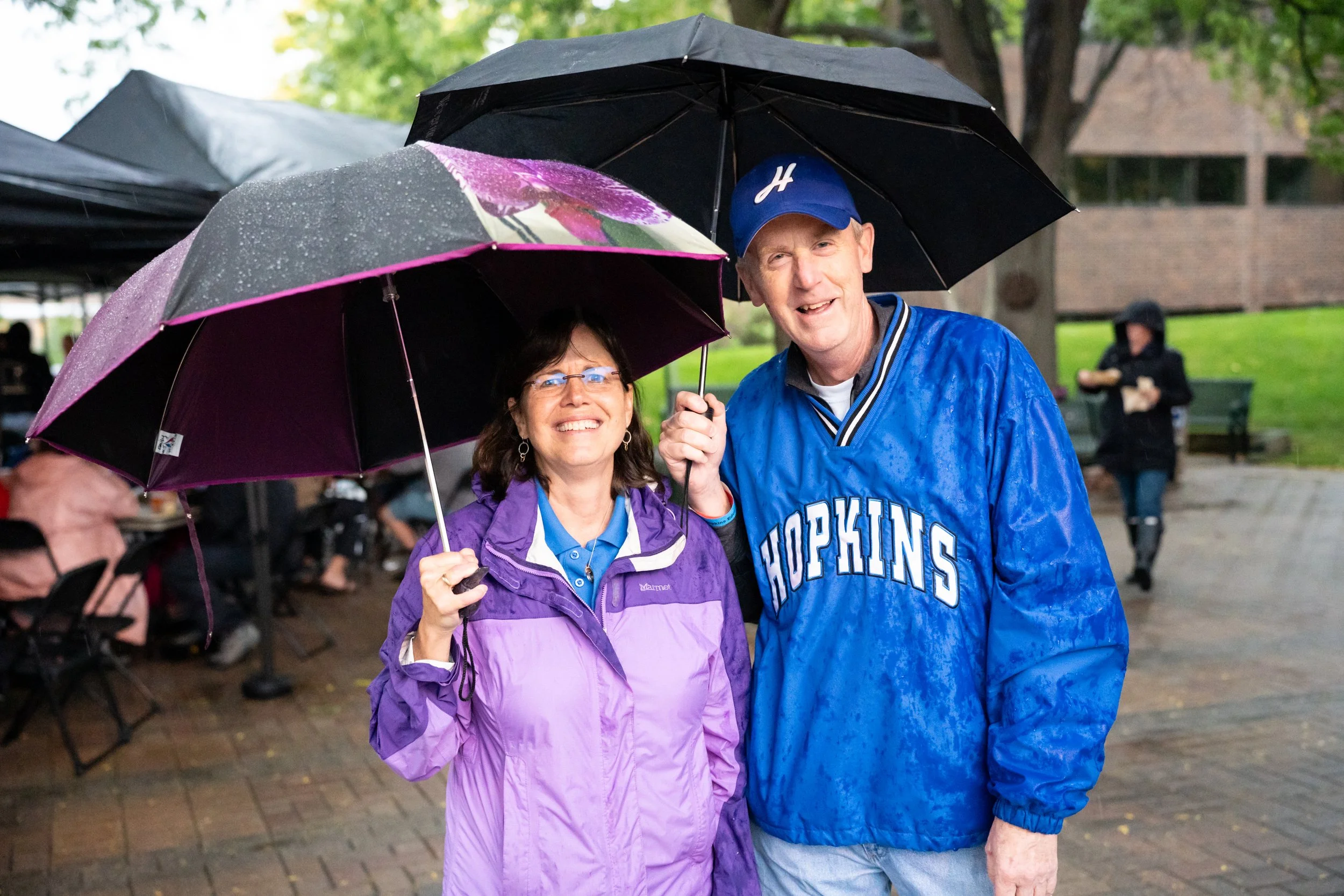 Two white adults stand outside in the rain with umbrellas