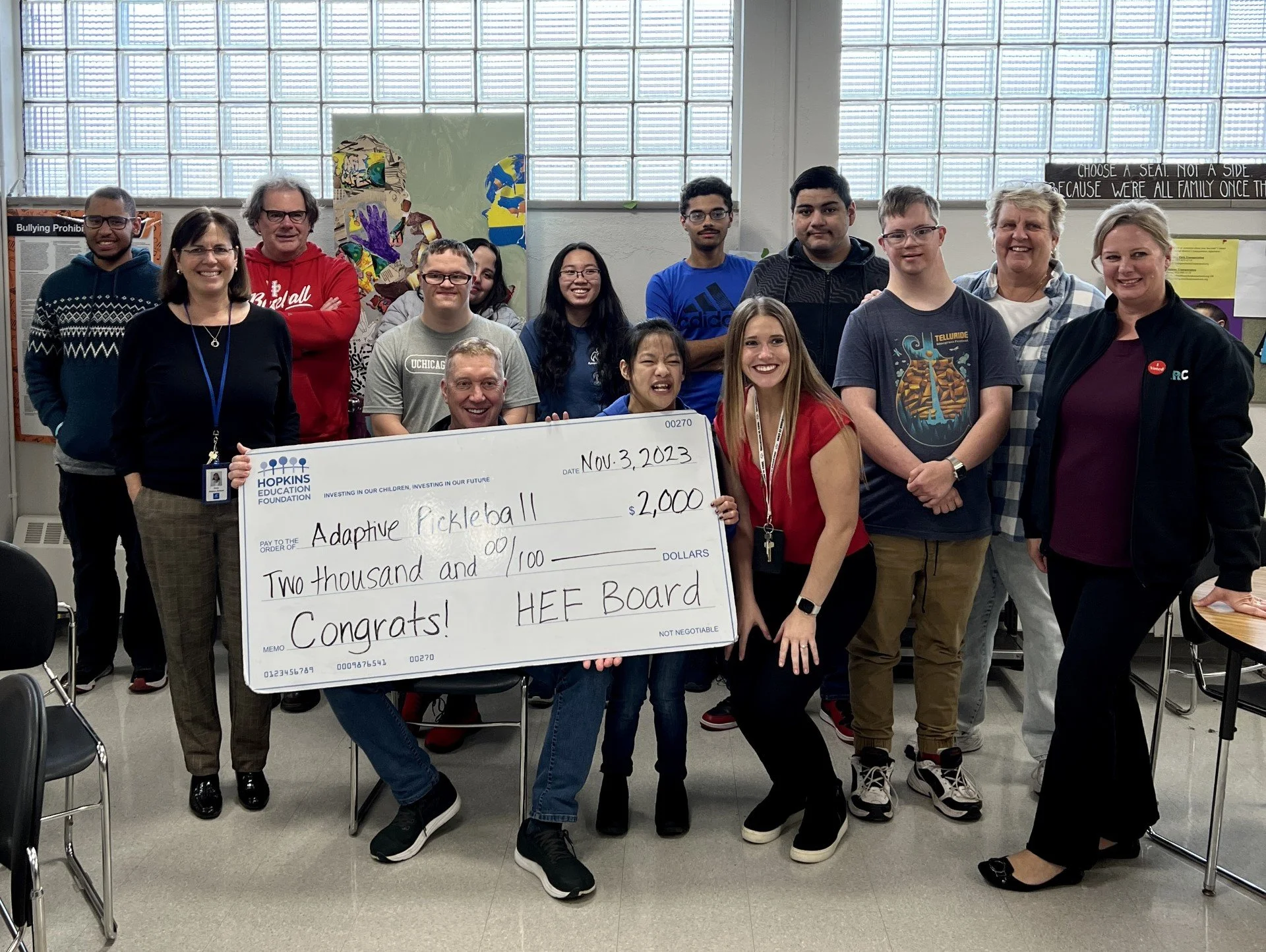 Group of secondary school students, representing a mix of genders and ethnicities, stand with teachers and board members with large grant check in foreground