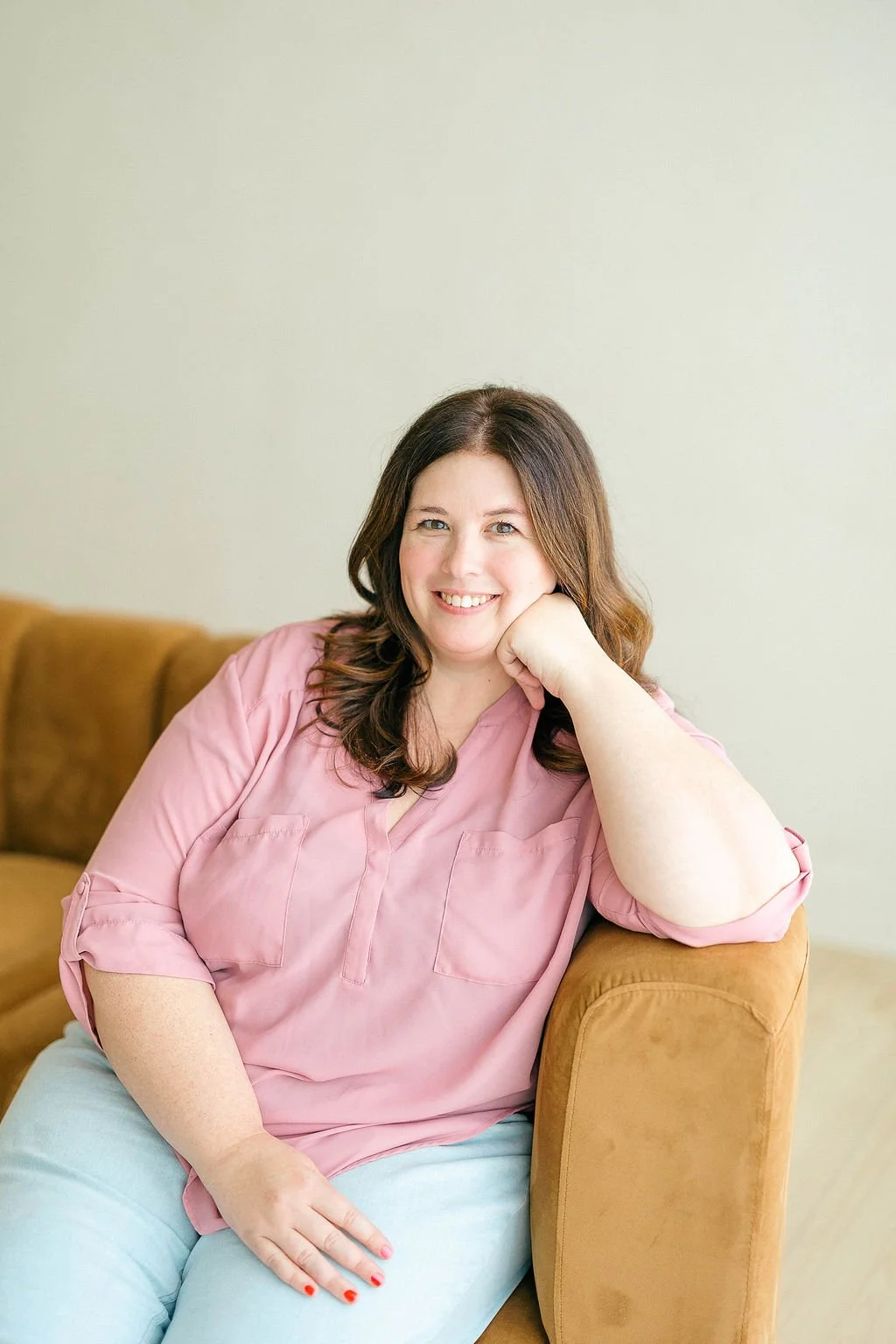 A smiling woman with brown hair in a pink blouse sitting on a brown sofa.