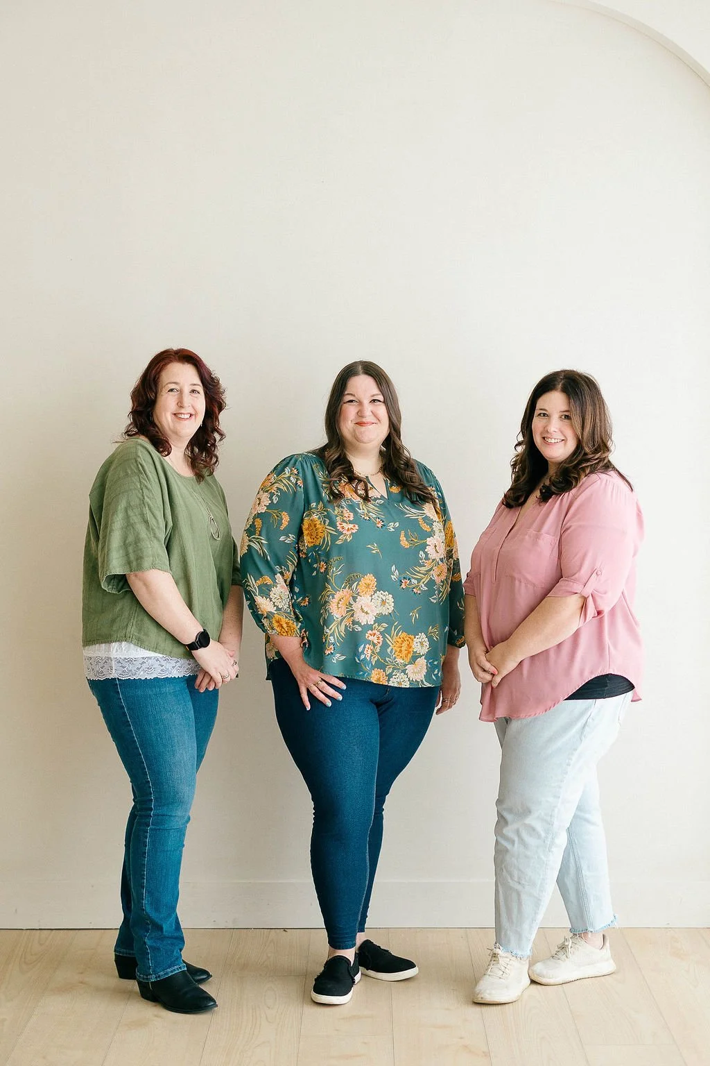 Three women standing side by side against a plain off-white wall, smiling at the camera.
