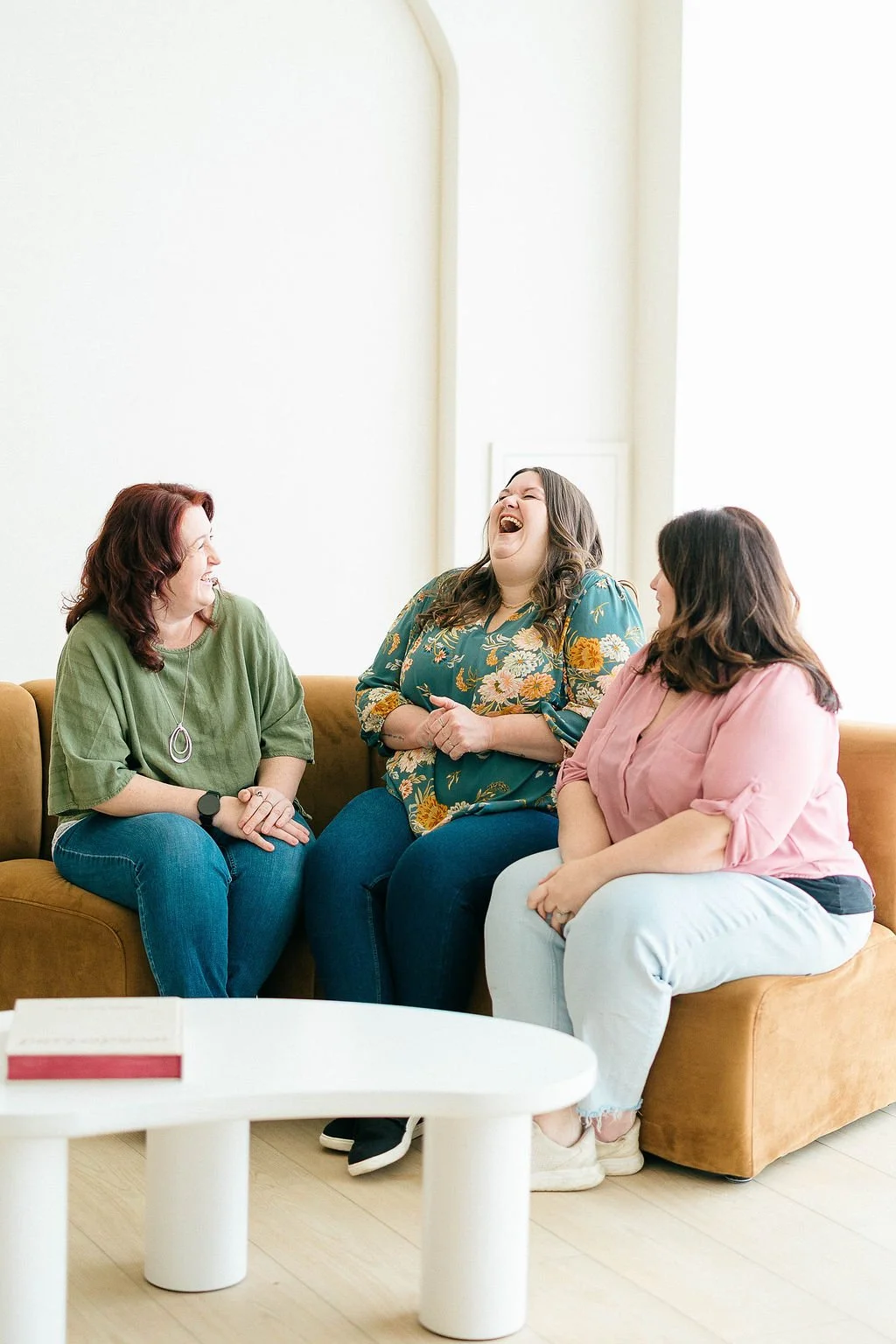 Three women sitting on a sofa, laughing and having a conversation in a bright room.