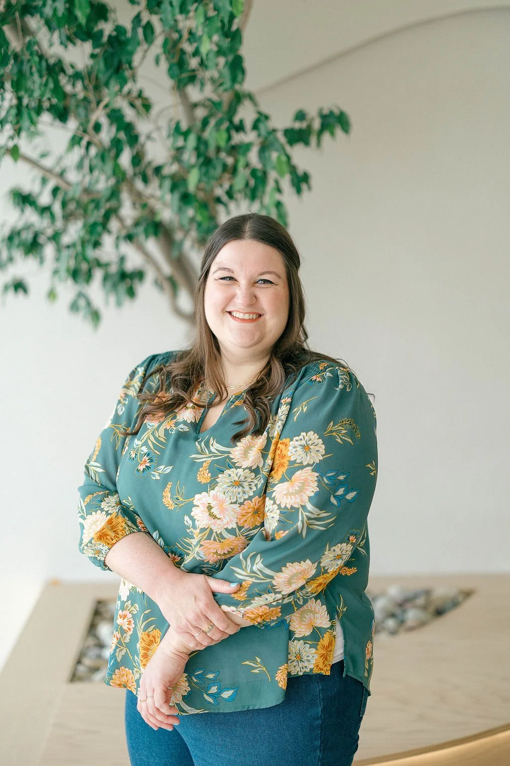 A woman with long brown hair smiling and wearing a floral teal blouse stands in front of a green leafy plant, indoors.