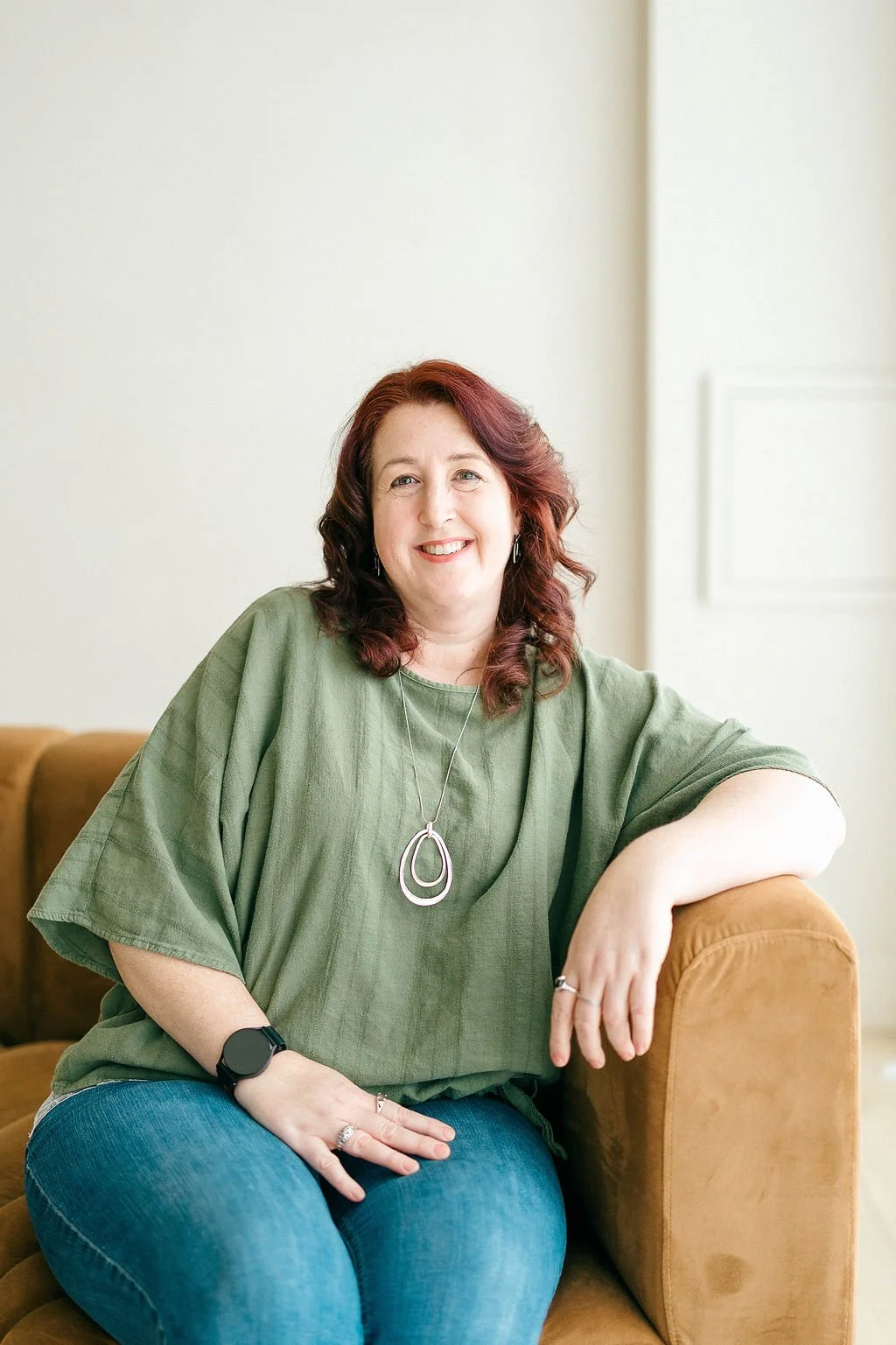 A smiling woman with red hair sitting on a brown couch in a light-colored room, wearing a green top, blue jeans, a large necklace, rings, an earring, and a smartwatch.
