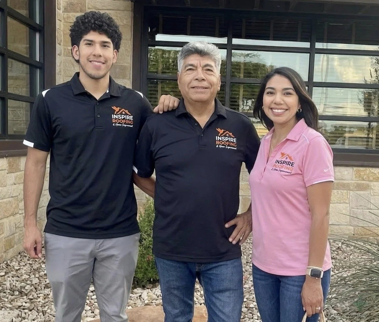 Three people standing outdoors in front of a building with large windows, wearing Inspire Roofing polo shirts, smiling at the camera.