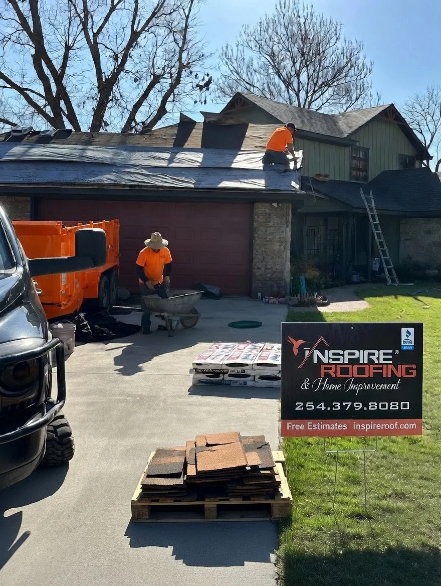 Two workers are replacing the roof on a house, one is on the roof installing new shingles, the other is on the ground with a wheelbarrow near a sign for Inspire Roofing.