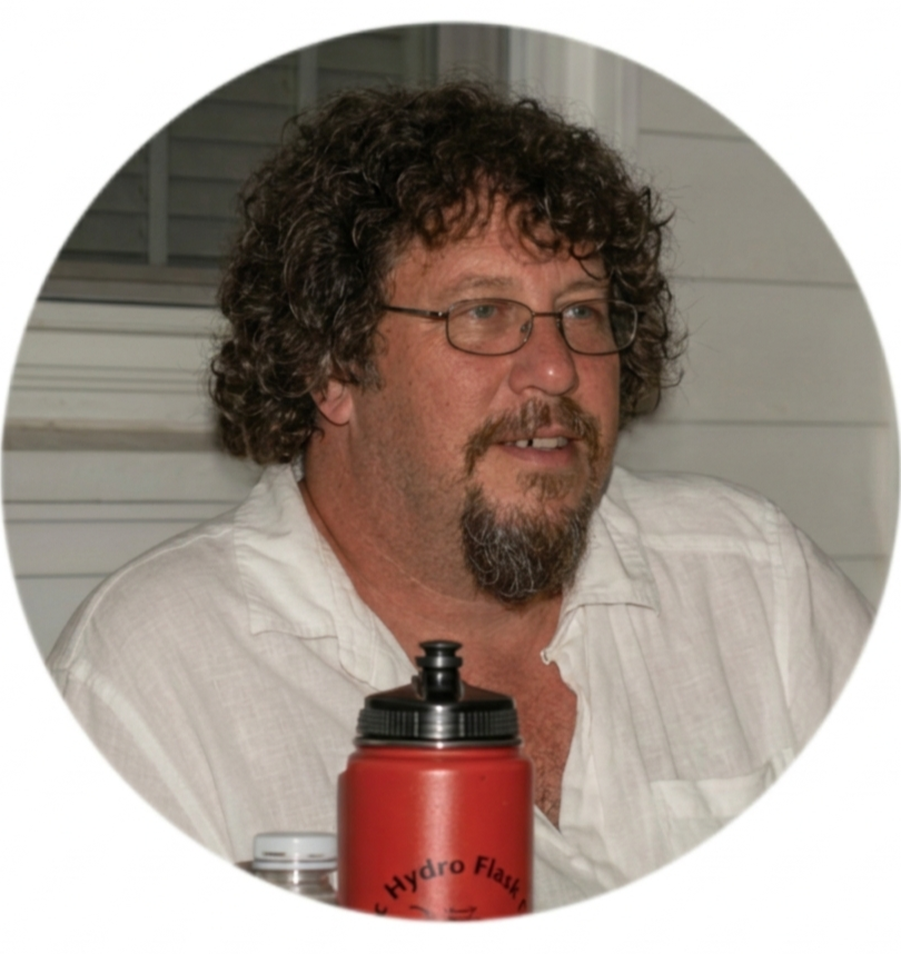 A man with curly hair, glasses, and a beard, wearing a white shirt, sitting indoors with a red Hydro Flask bottle in front of him.