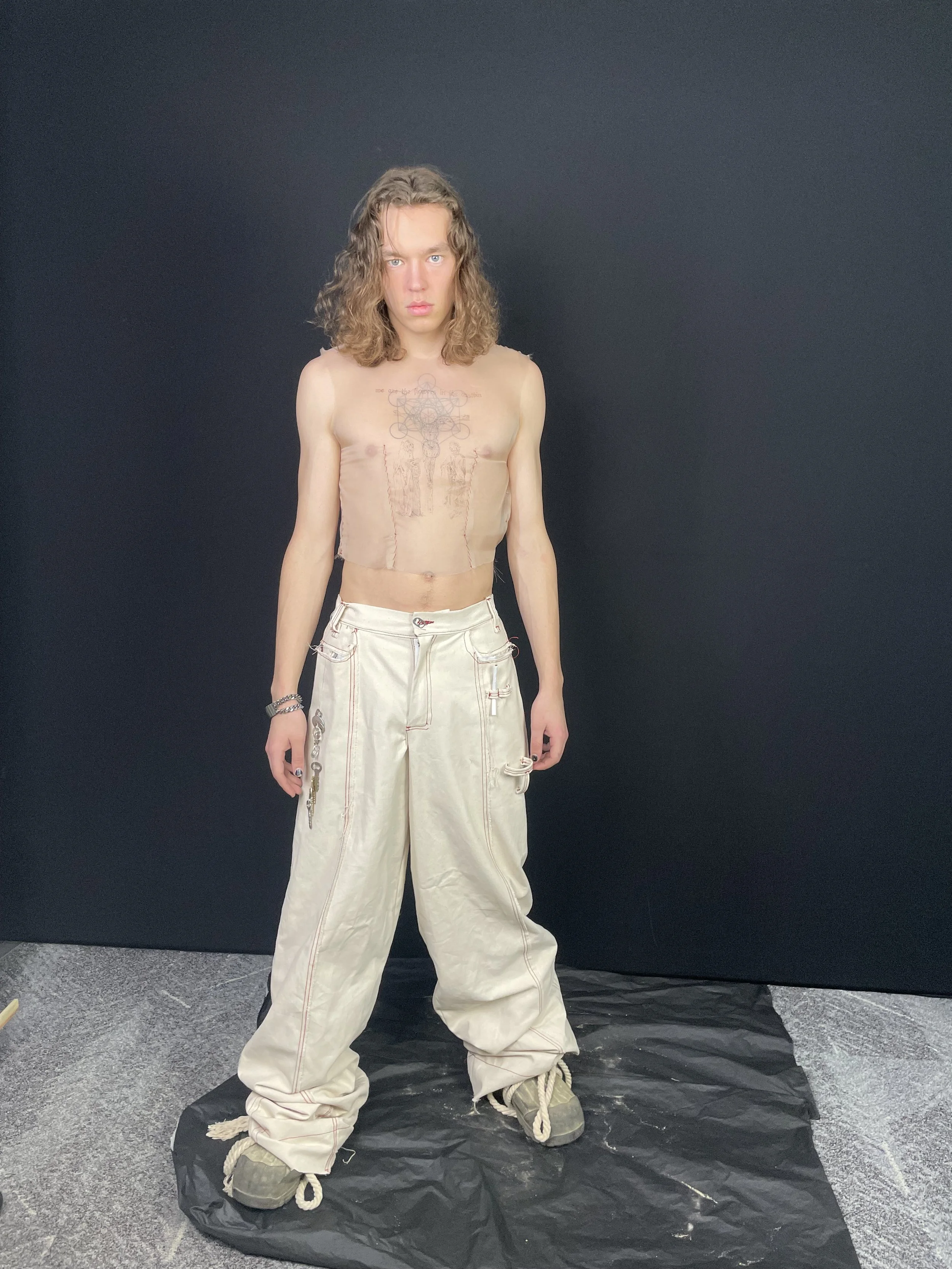 A young man with long, curly hair standing against a black background, wearing baggy beige pants and no shirt, showing tattoos on his chest and abdomen. conor o'driscoll