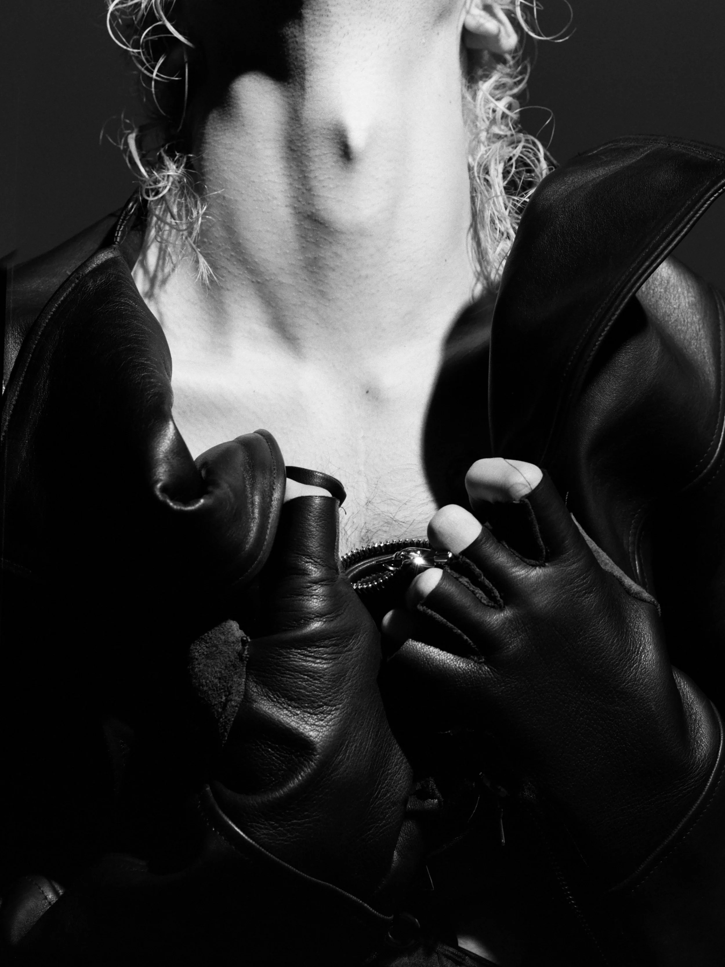 Close-up black-and-white photo of a person with curly hair, wearing leather gloves and a leather jacket, holding the zipper of the jacket, with the lower face and neck visible. conor o'driscoll