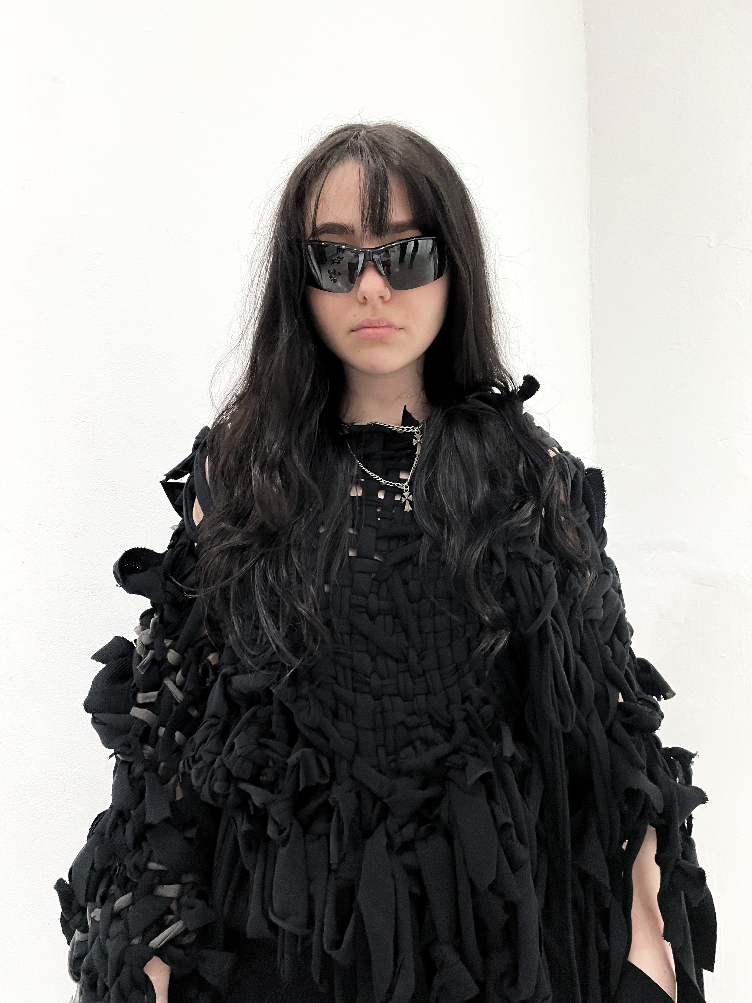 A young woman with long, dark, wavy hair wearing black sunglasses, a black distressed outfit with various straps and fabric knots, and layered silver necklaces, standing against a plain white wall. conor o'driscoll
