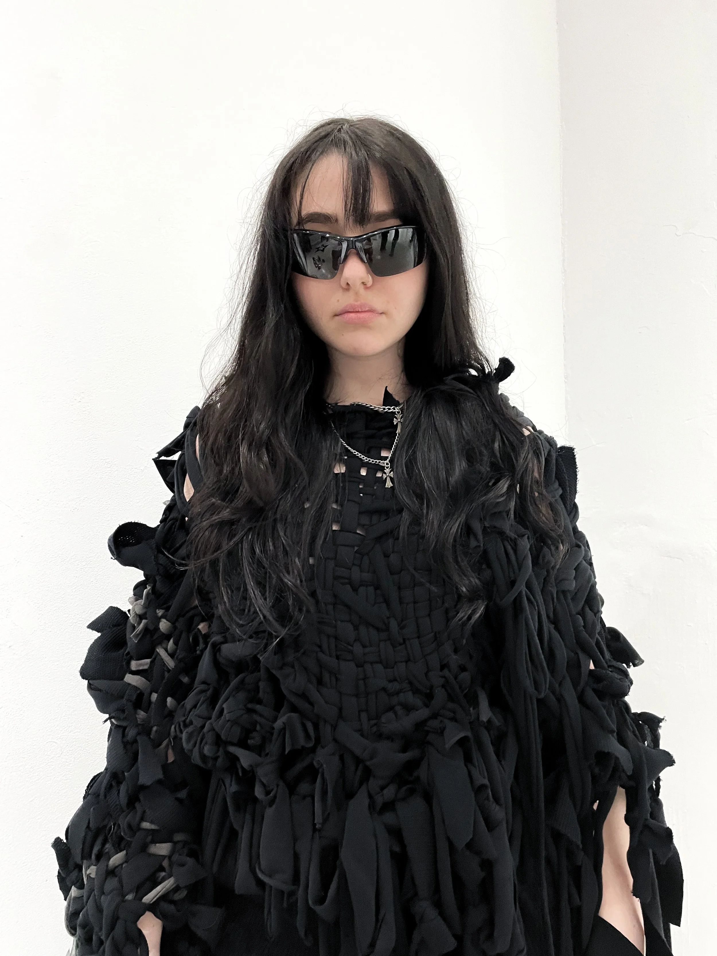skye wilson long, dark, wavy hair wearing black sunglasses, a black distressed outfit with various straps and fabric knots, and layered silver necklaces, standing against a plain white wall. conor o'driscoll