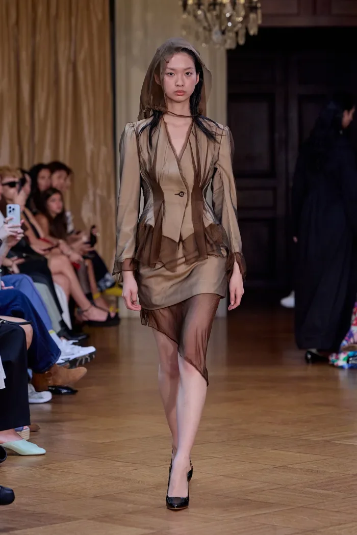 CONOR ODRISCOLL FOR VETTESE. S26 NYFW Fashion runway with model in beige and brown layered outfit, black heels, and an audience in background.