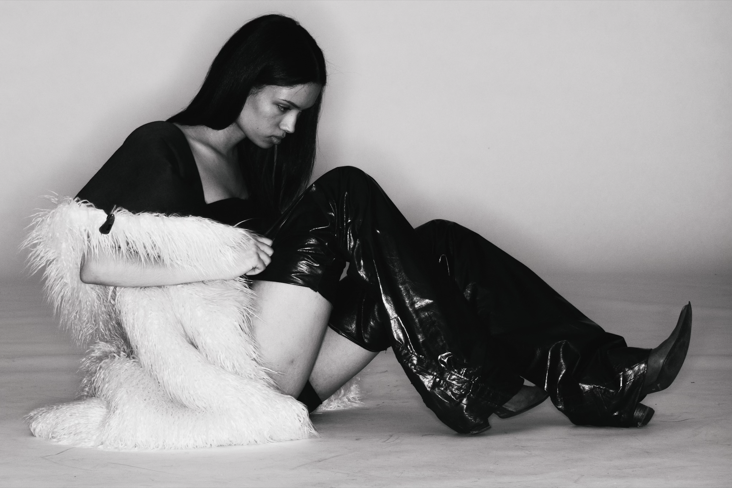 A young woman with long dark hair, wearing a black top, shiny black pants, and boots, sitting on the floor against a plain background, with a white faux fur coat draped over her arm, looking downward. conor o'driscoll NICO DANIELS