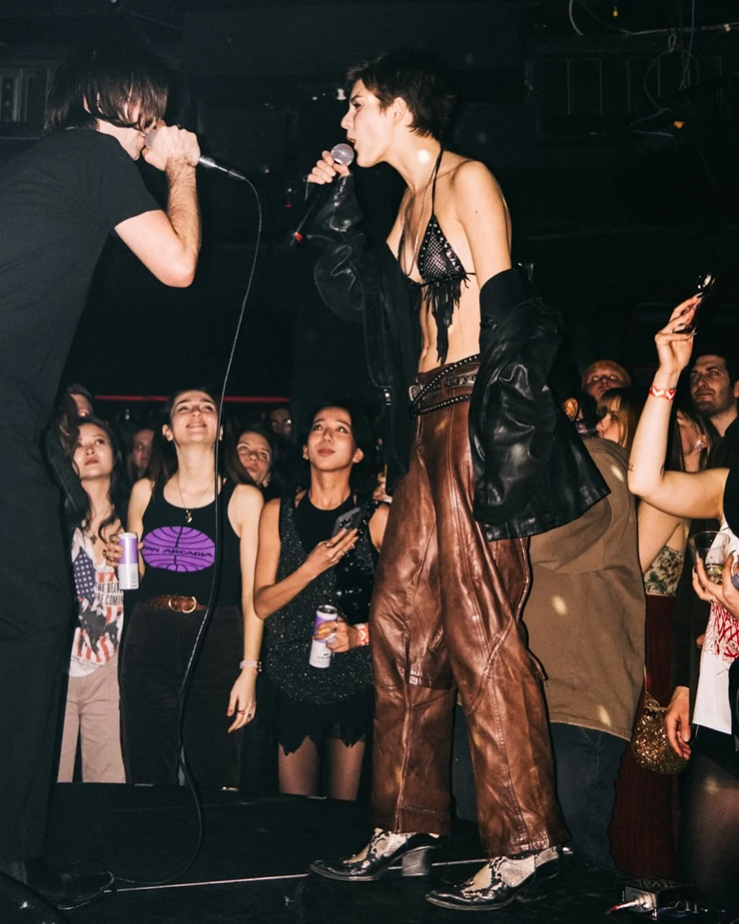 A woman singing into a microphone on stage at a concert with a crowd watching, some holding drinks or phones, in a dark indoor venue. by conor o'driscoll