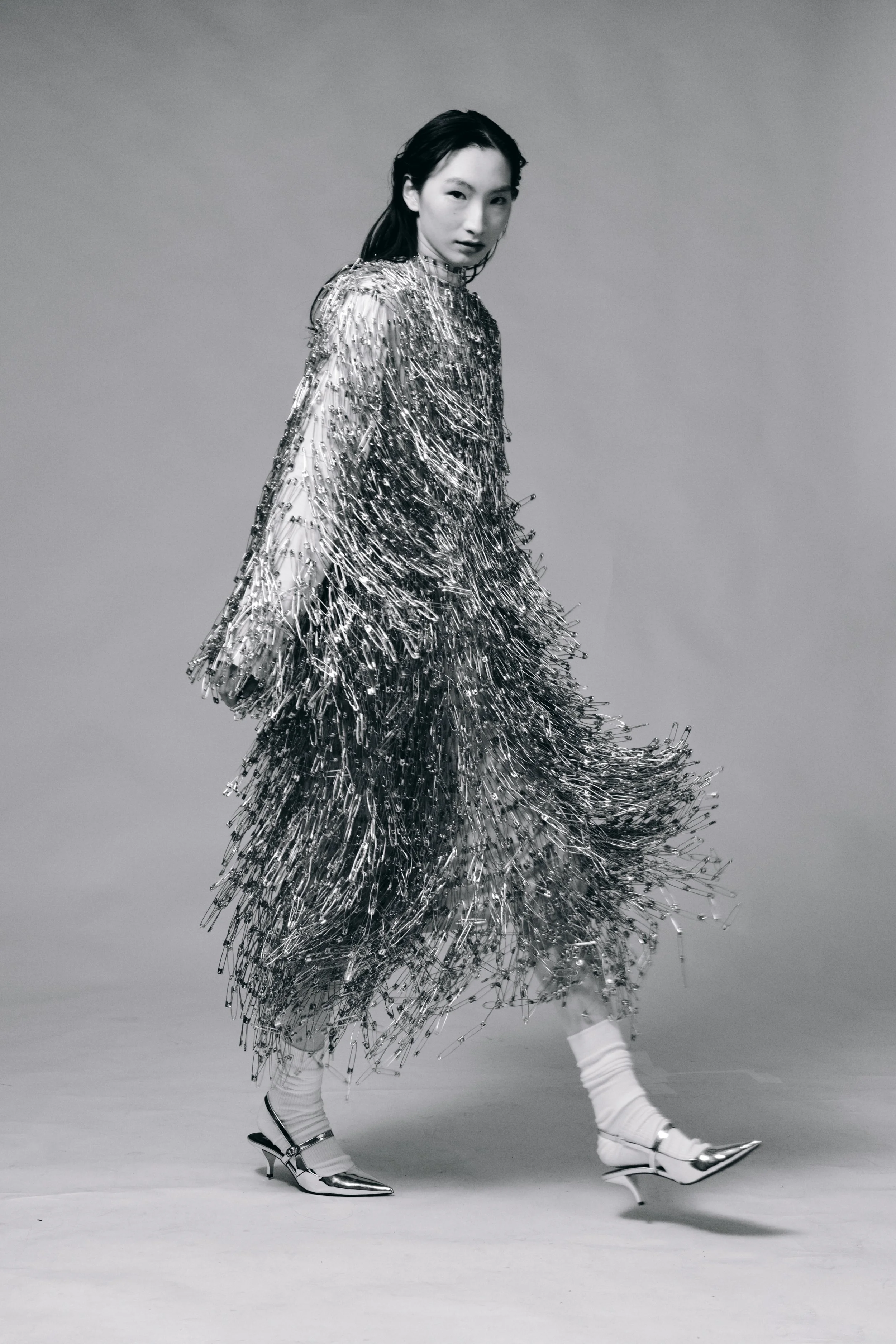 Fashion model wearing a metallic, fringed dress with high-heeled shoes and socks, posing in a studio with a plain backdrop. conor o'driscoll