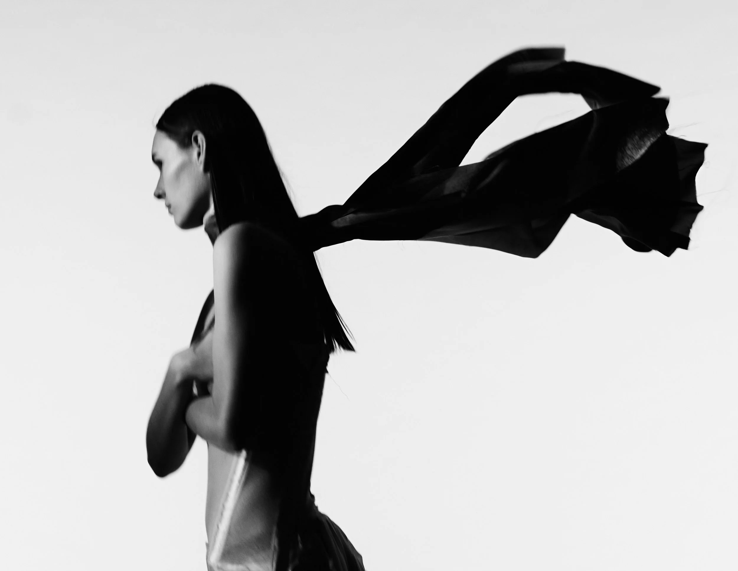 Black and white silhouette of a woman with long hair, her arm crossed over her chest, and a flowing scarf or fabric extending behind her.conor o'driscoll