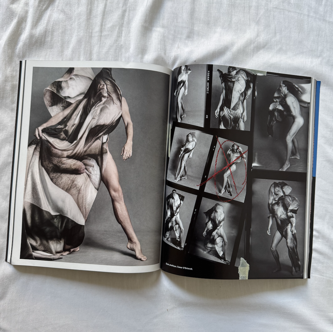 An open art book showing a large photograph of a model with a sheet of fabric covering their body, surrounded by smaller black-and-white photos of the same model in various poses, some with red crosses and markings. Conor O'Driscoll