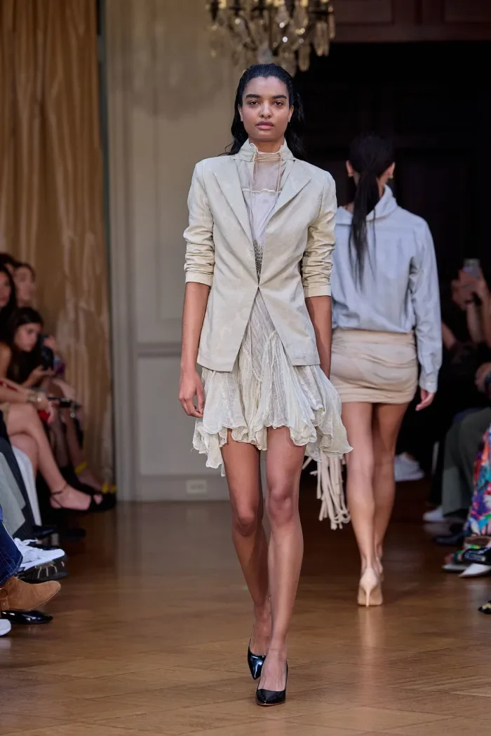 CONOR ODRISCOLL FOR VETTESE. S26 NYFW Model walking down runway in beige blazer and light-colored ruffled dress at fashion show.