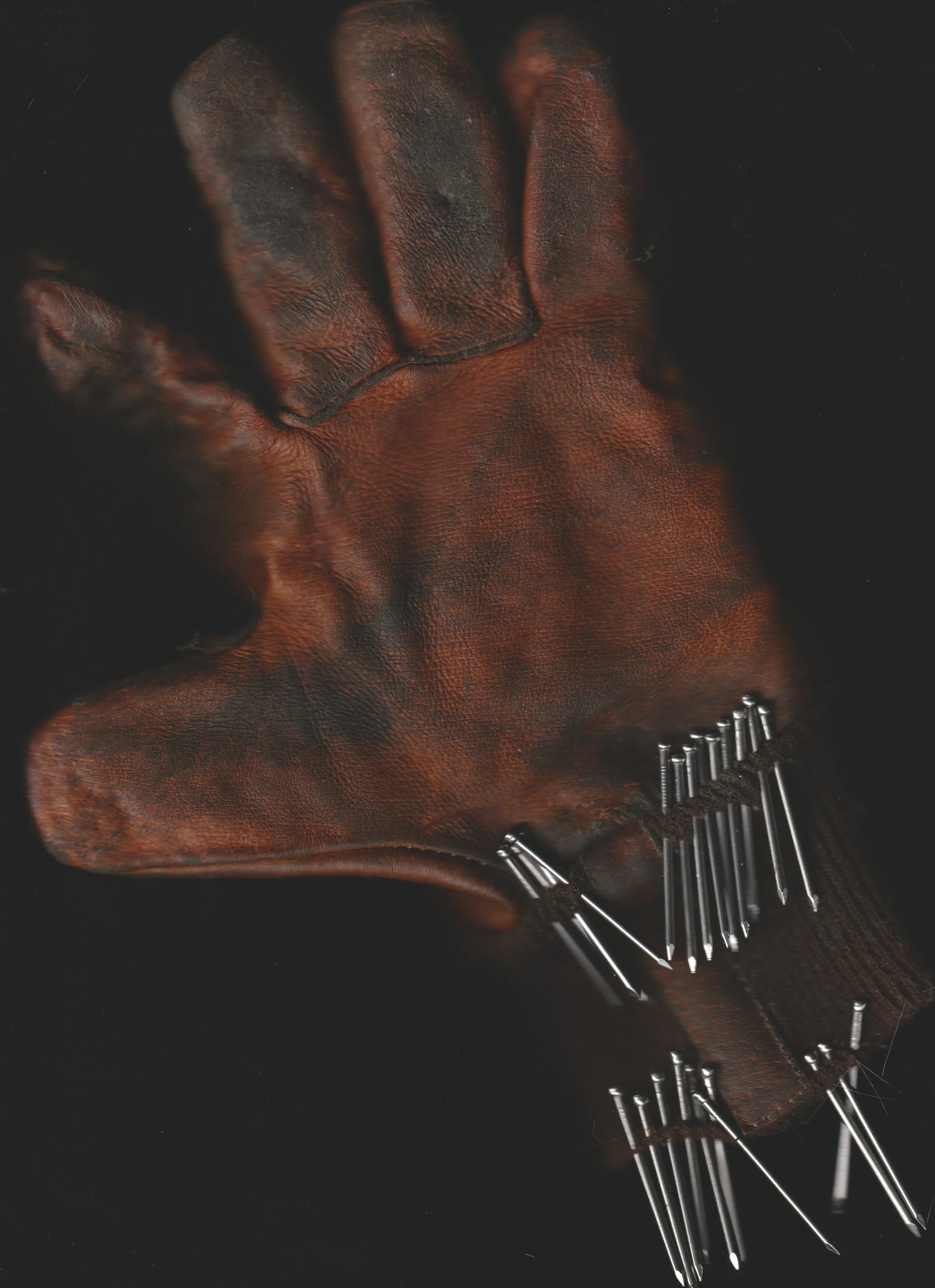 A worn, brown leather work glove with numerous sewing pins attached to its cuff area. conor o'driscoll