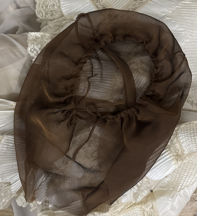 CONOR ODRISCOLL FOR VETTESE. S26 NYFW A brown, sheer, circular cloth with a tie in the middle, placed on a bed with white lace and striped bedding.