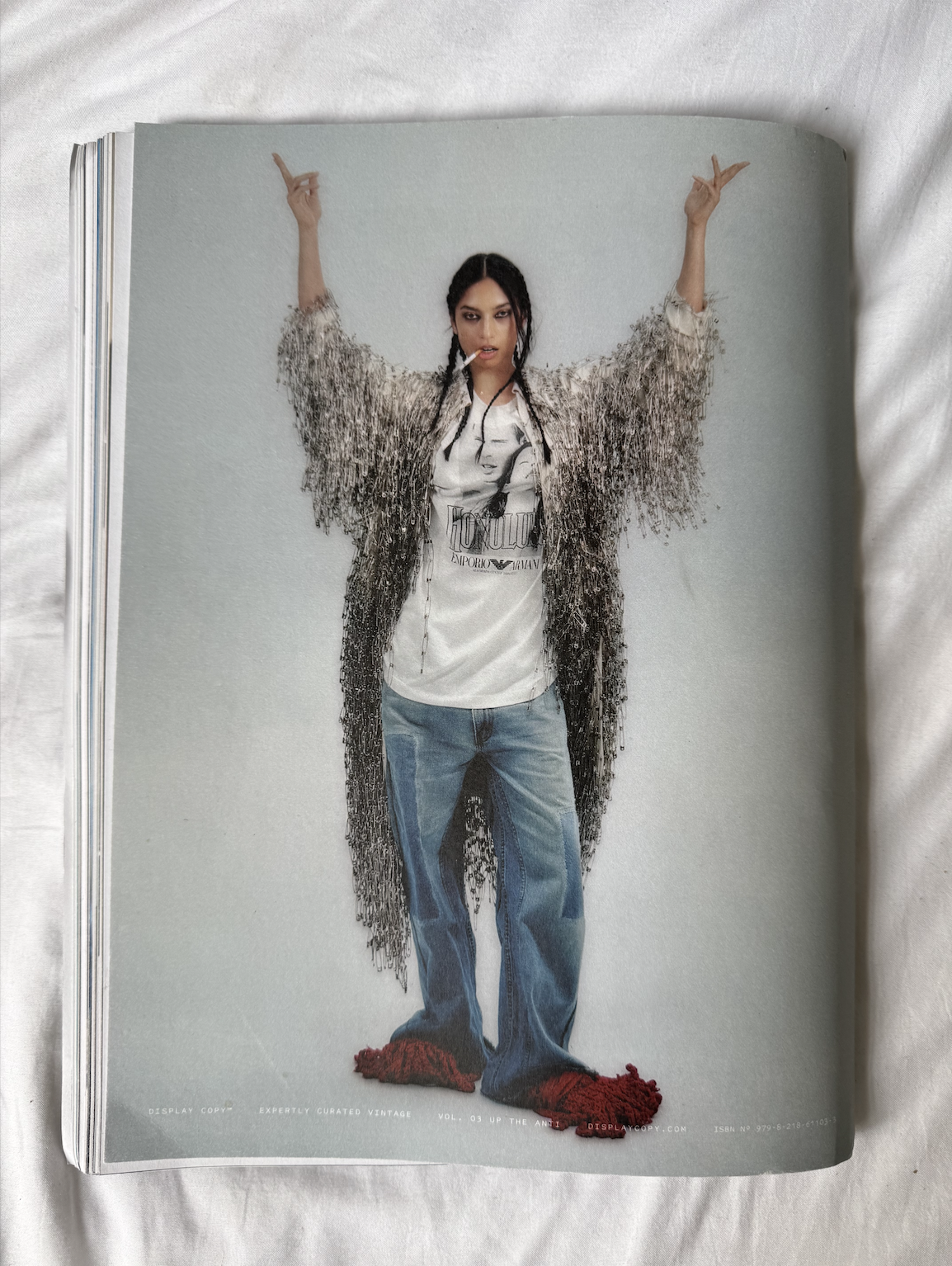 Fashion magazine page featuring a woman with dark hair in braids, wearing a graphic T-shirt and jeans, draped in a fringed safety pin coat, standing with arms raised and a cigarette in her mouth.  CONOR O'DRISCOLL
