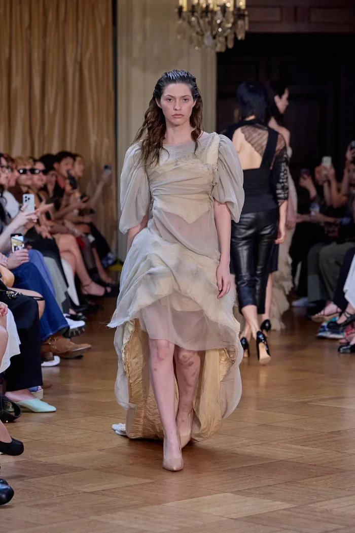 CONOR ODRISCOLL FOR VETTESE. S26 NYFW Model walking on runway in beige and cream layered dress with puffed sleeves, audience seated on both sides