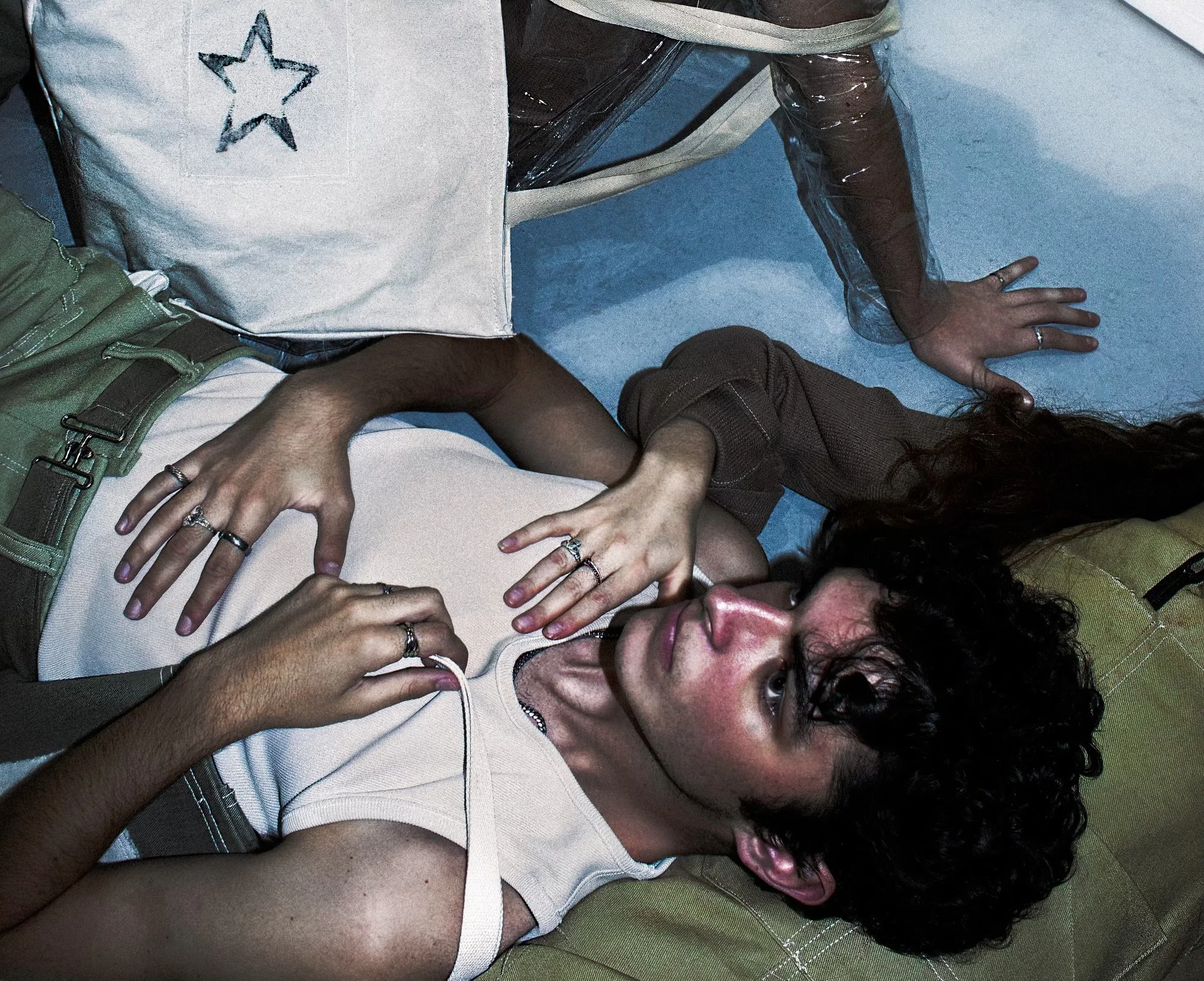 Two young women lying on the floor, one on her back and the other on her stomach, surrounded by bags and furniture, with the woman on her back looking up at the camera. conor o'driscoll