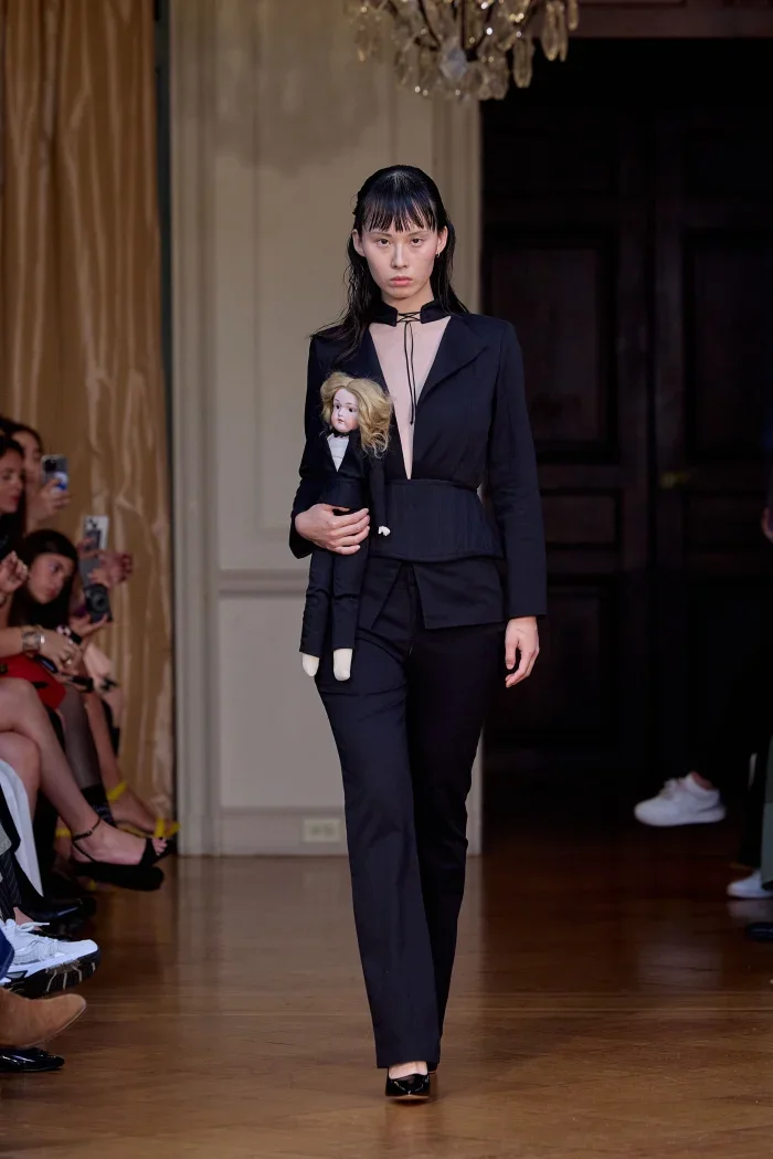 CONOR ODRISCOLL FOR VETTESE. S26 NYFW Model walking down runway holding a doll dressed in black at a fashion show.