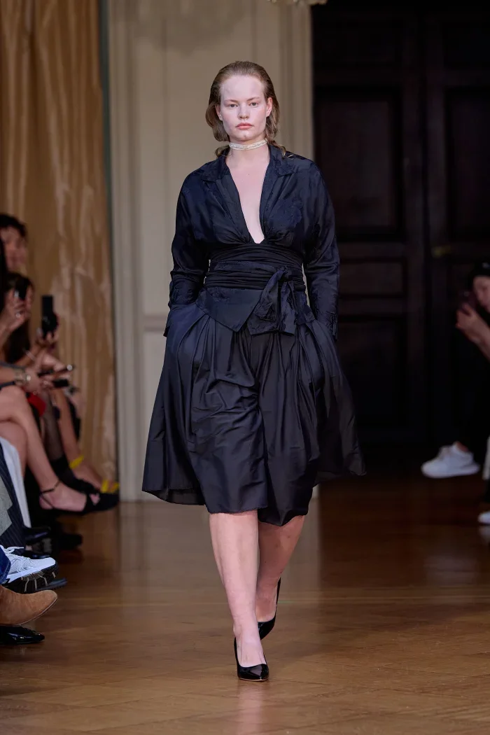 CONOR ODRISCOLL FOR VETTESE. S26 NYFW A female model walking on a runway in a black layered dress with a deep V-neck and a belt around her waist, paired with black high heels.