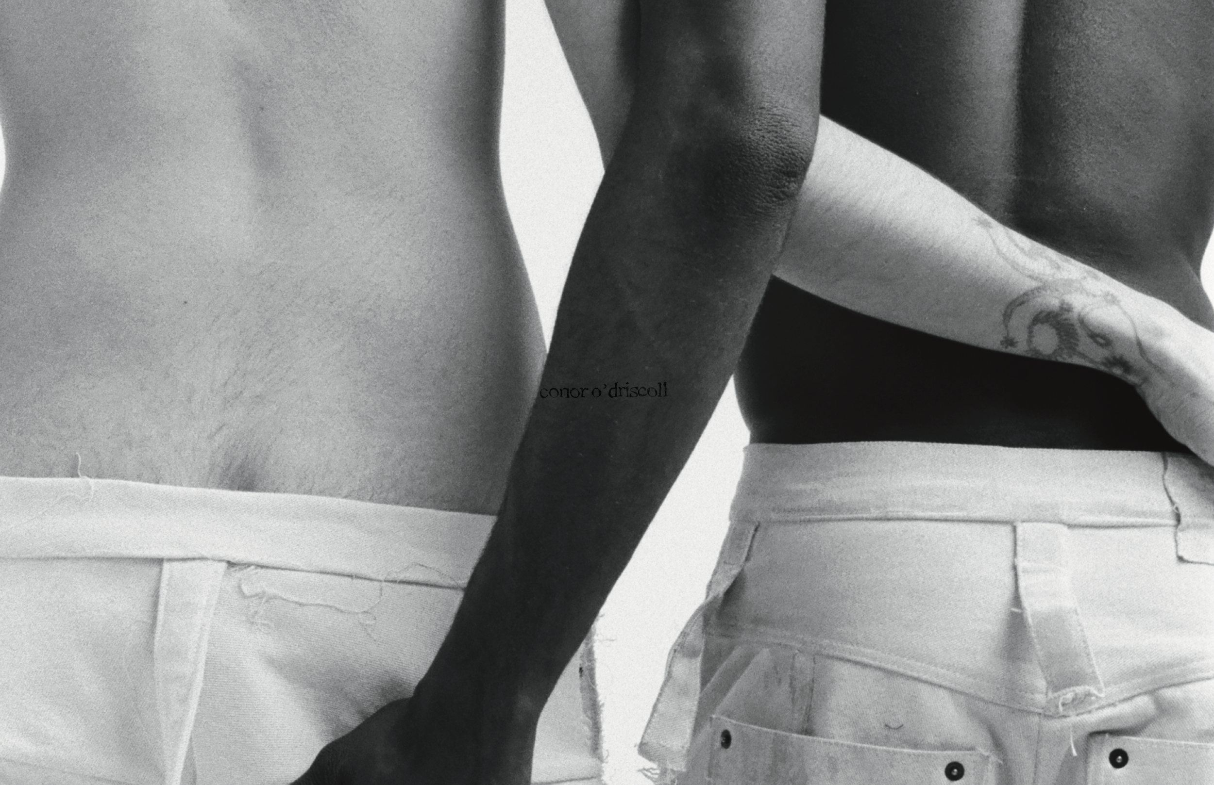 Close-up black and white photograph of two shirtless people with varying skin tones, wearing light-colored shorts. One person's torso and arm are visible, with a tattoo on their side reading 'conor o'driscoll'.