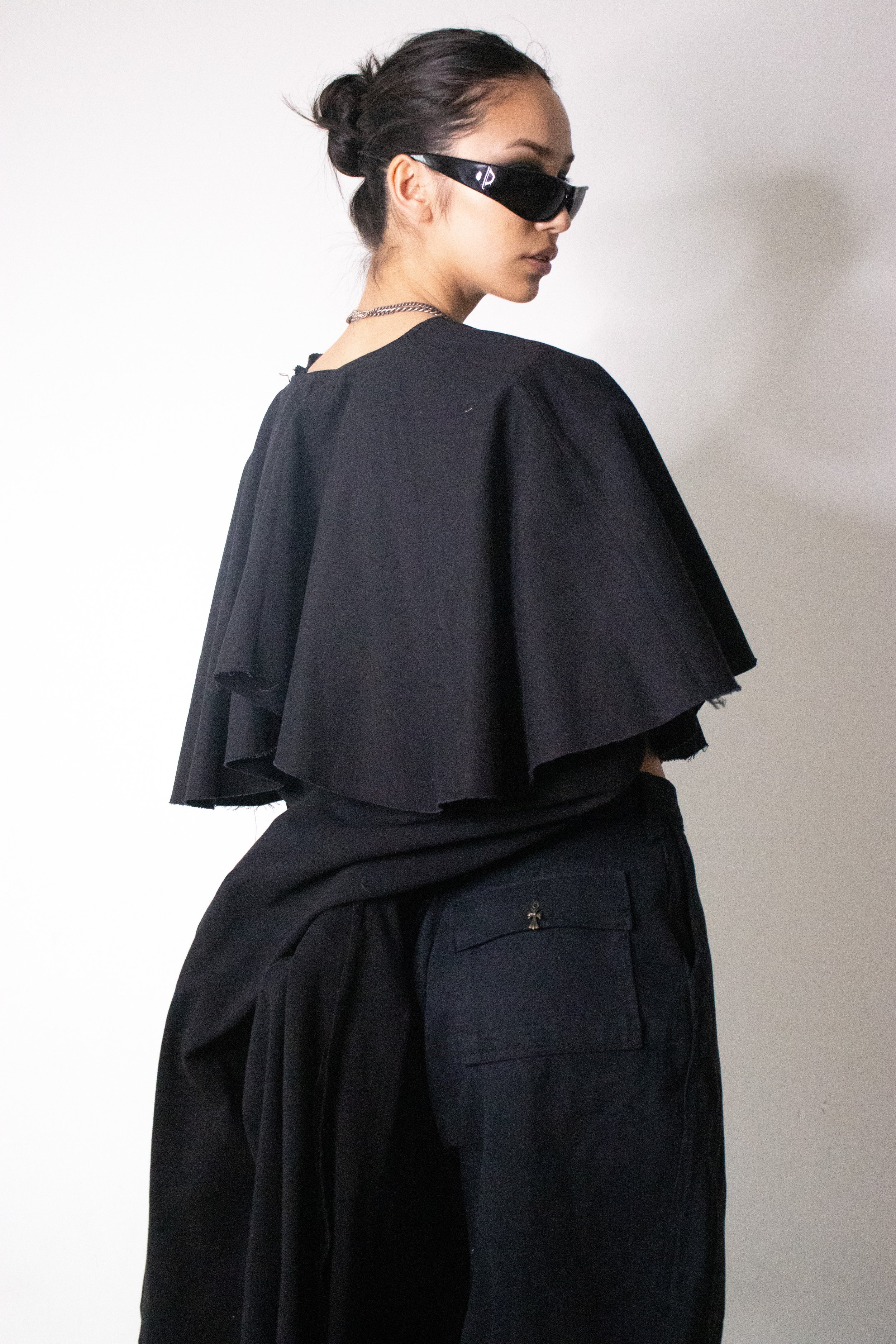 A woman with dark hair styled in a bun, wearing black sunglasses, a black top with ruffled sleeves, and black pants standing against a light background. conor o'driscoll