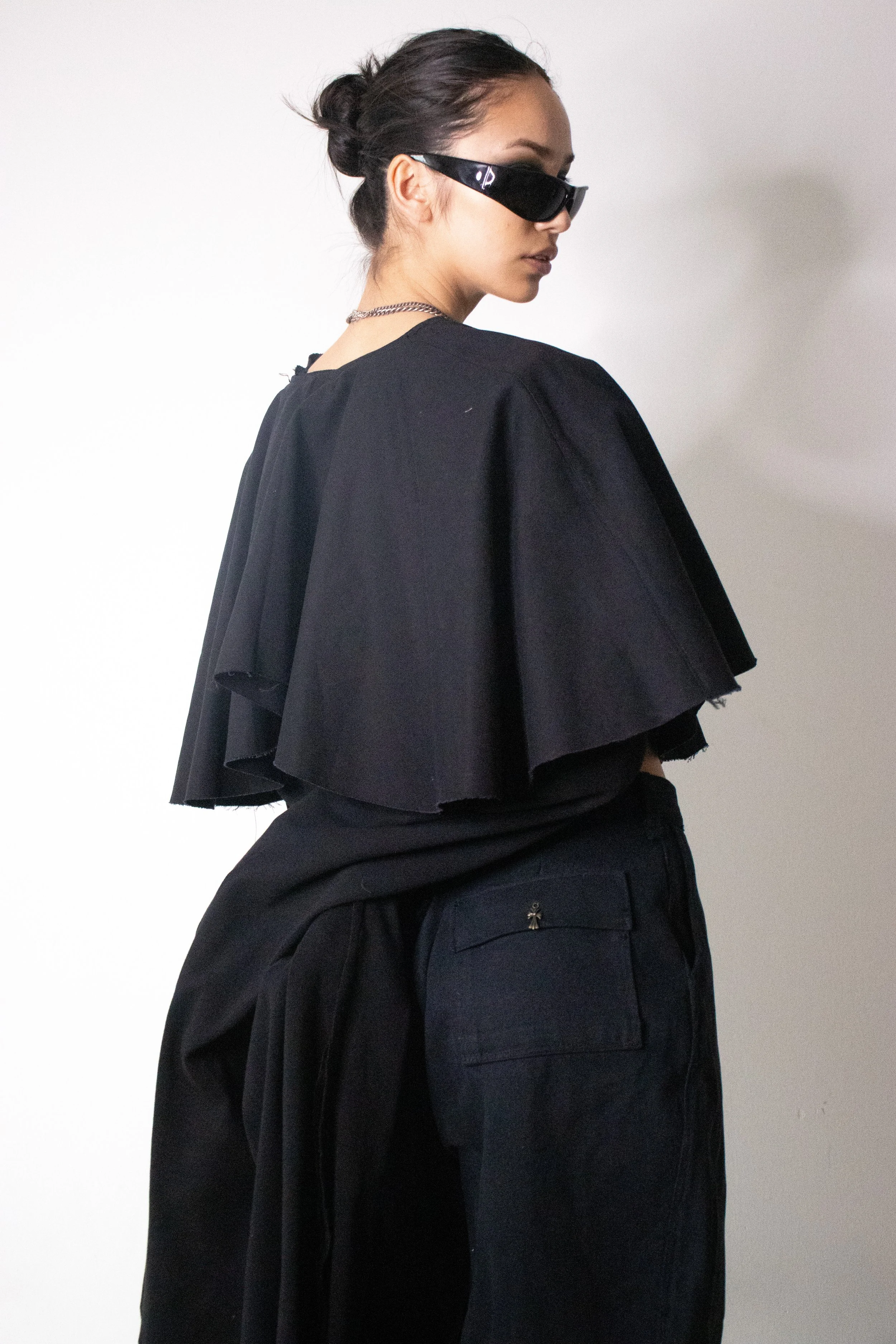 A woman with dark hair styled in a bun, wearing black sunglasses, a black top with ruffled sleeves, and black pants standing against a light background. conor o'driscoll